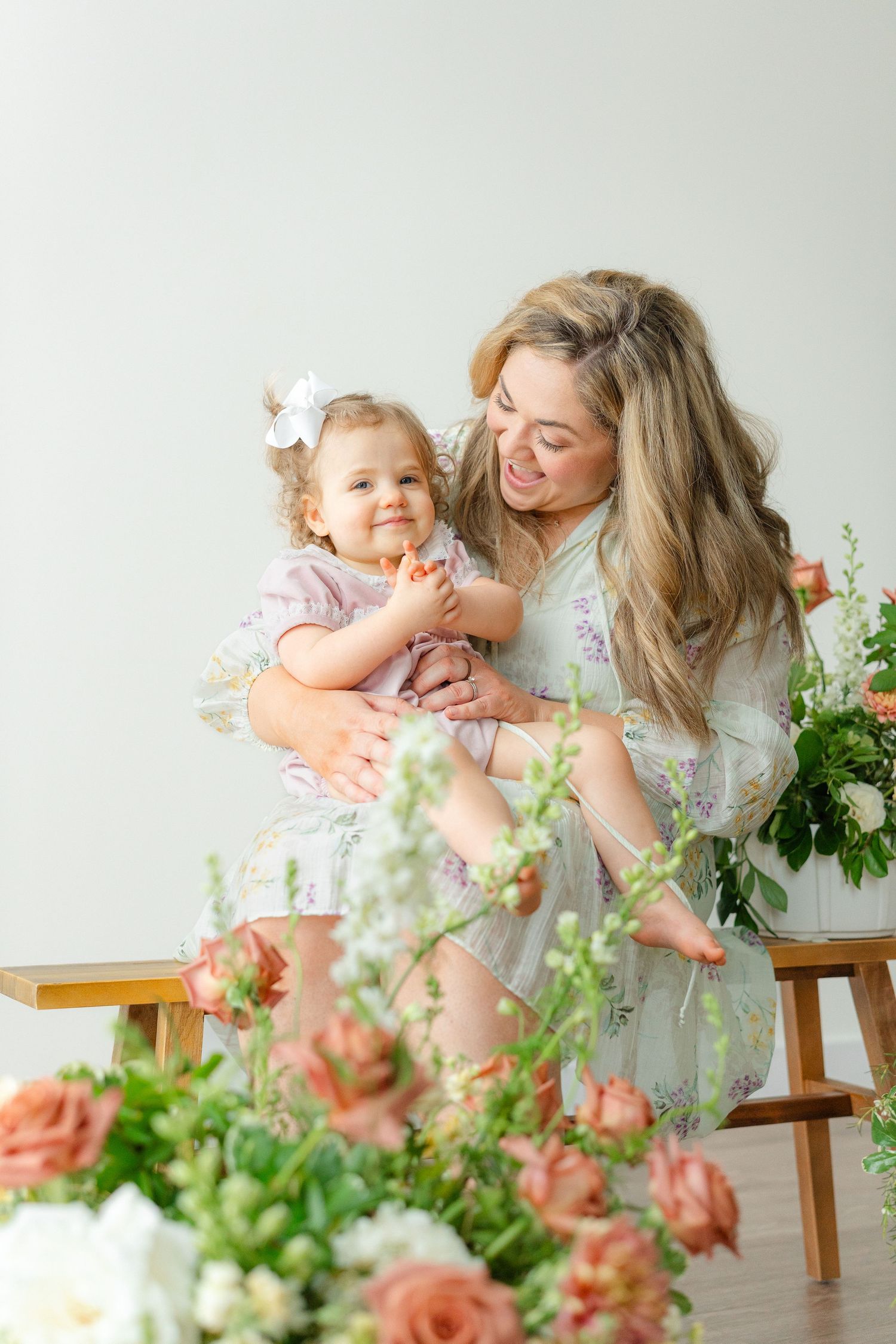 A smiling mother in a floral dress holds her laughing toddler daughter surrounded by fresh flowers.