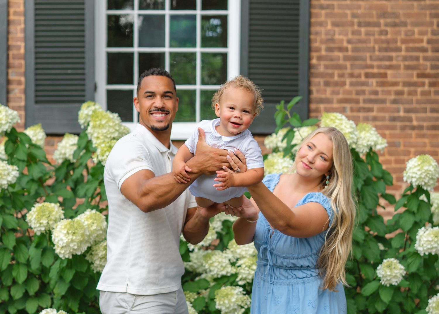 Happy family with parents holding smiling baby outdoors near white hydrangeas and brick home.