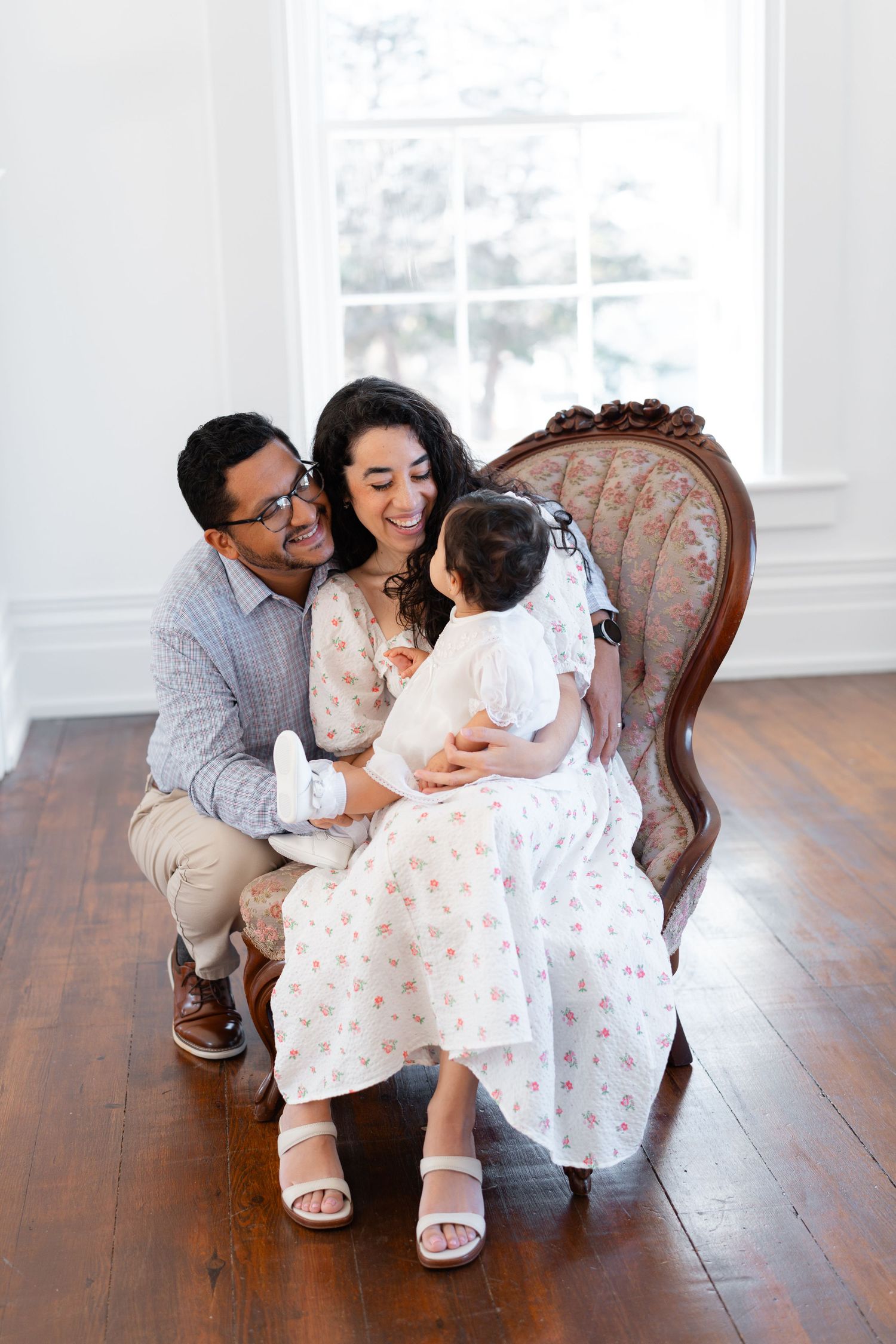 Smiling family with baby sitting on vintage floral chair in bright white room with hardwood floors.
