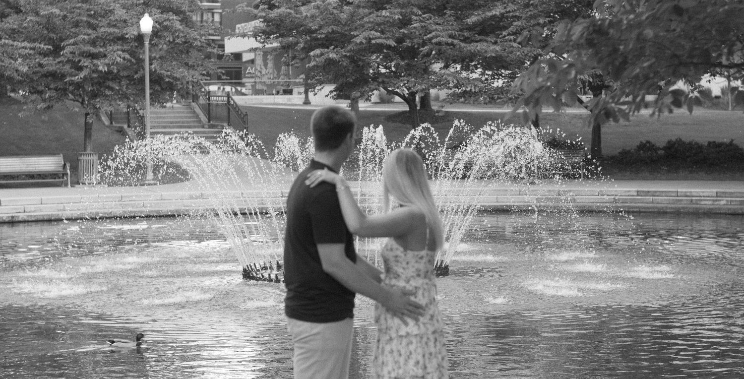 Black and white photo of a couple embracing in front of a decorative outdoor fountain.