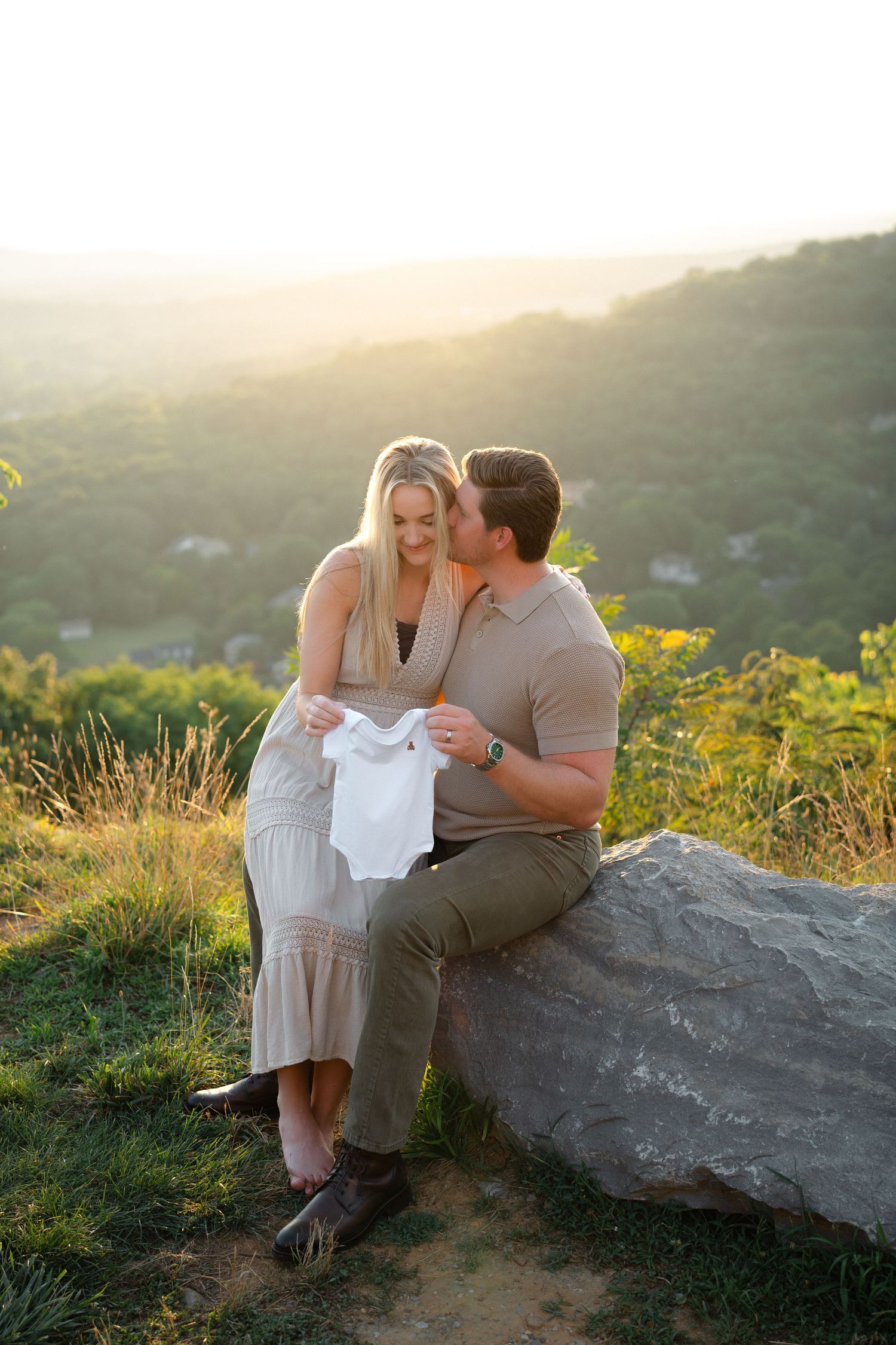 Couple sitting on a rock at golden hour holding a white baby onesie, announcing pregnancy with scenic hills behind.