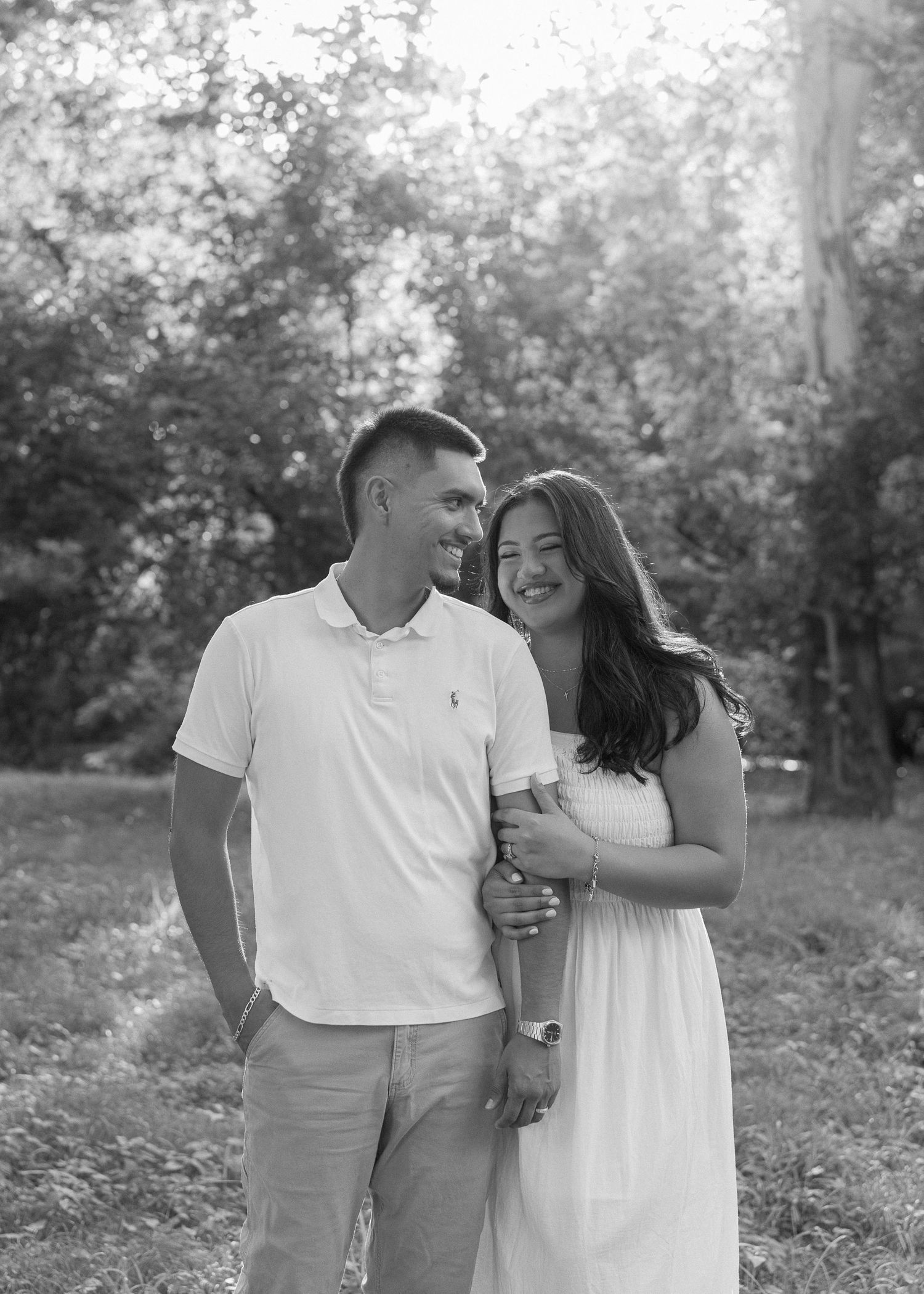 Black and white photo of a smiling couple embracing outdoors among lush trees in a park setting.