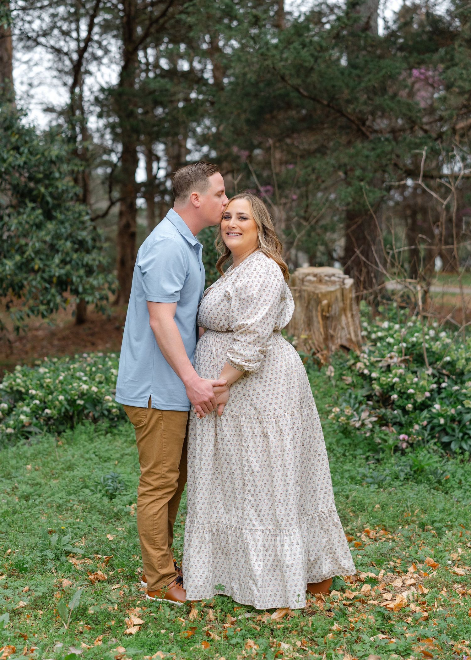 A pregnant woman in a floral maxi dress smiles as her partner kisses her head in a lush garden setting.