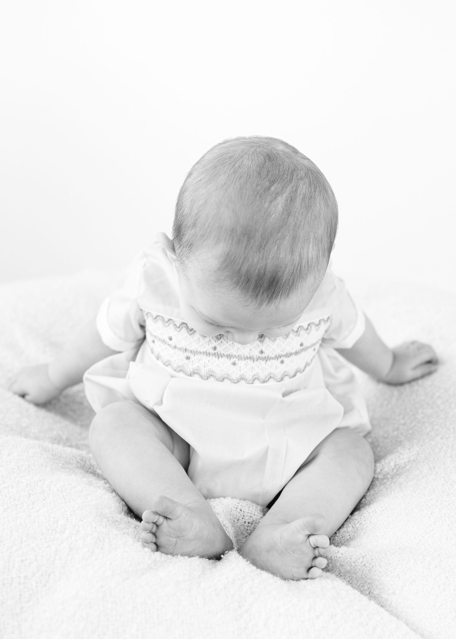 Black and white photo of baby sitting upright on soft blanket, viewed from behind.