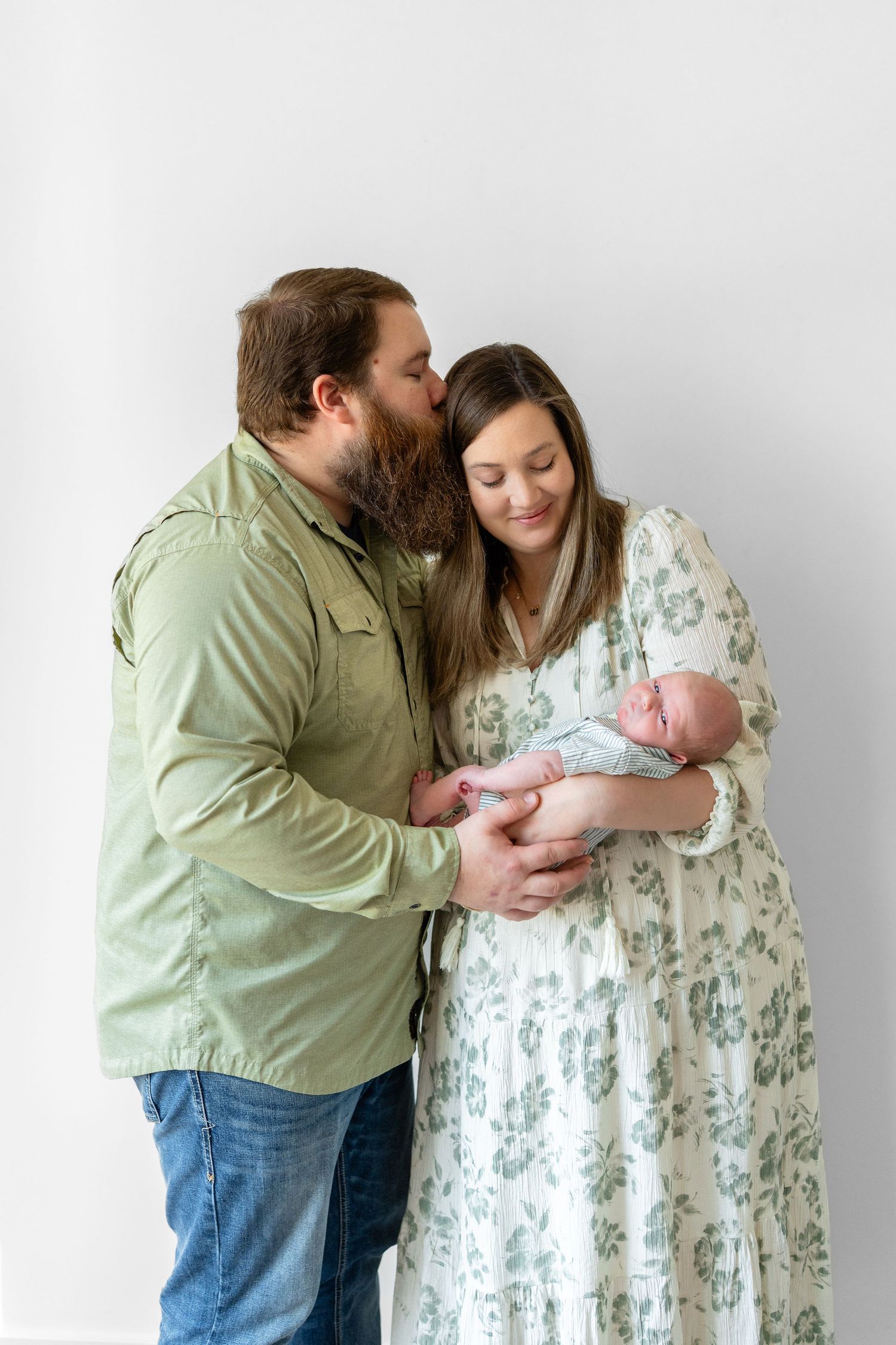 A bearded man in green kisses a woman in floral dress as she cradles their newborn baby against a white background.