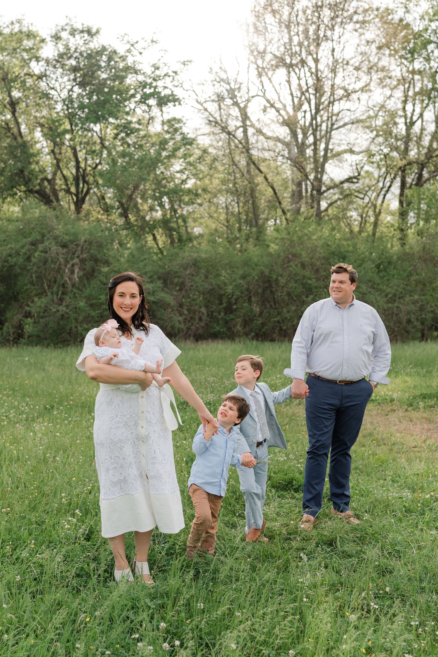 Family of five poses in a lush green meadow, mother holding newborn while two boys play nearby.