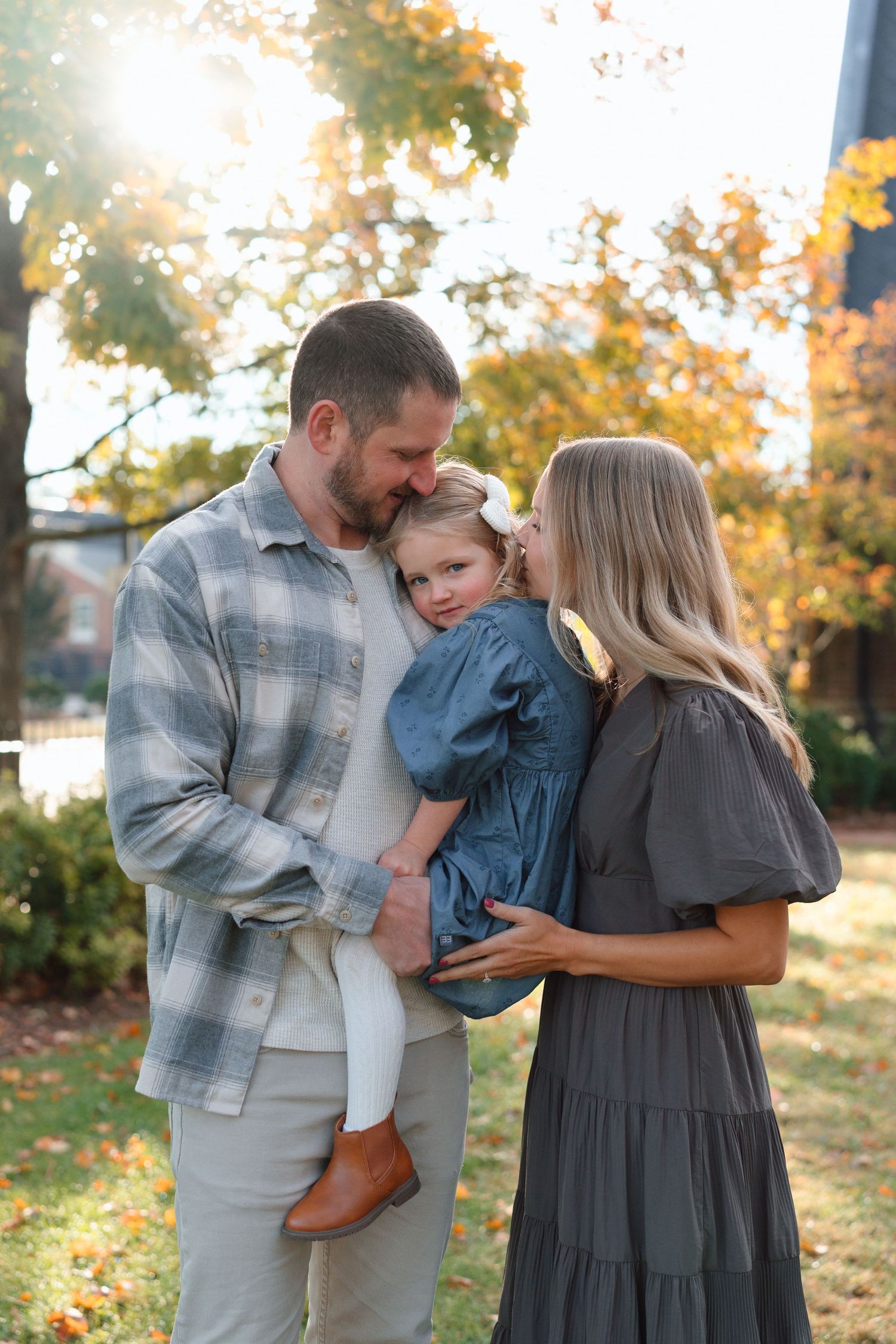 Parents lovingly kiss their young daughter outdoors in warm autumn sunlight.