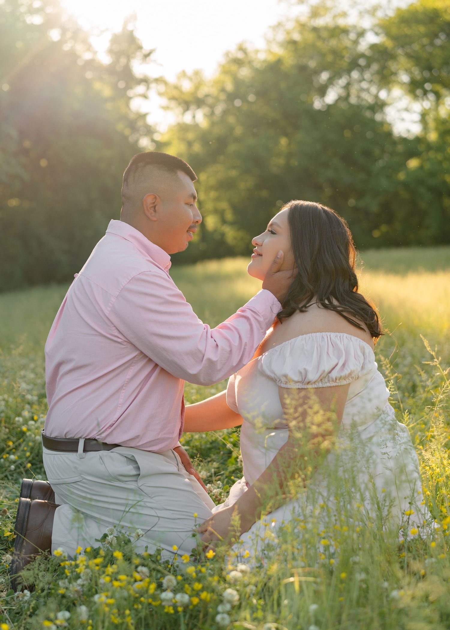 Couple sitting in a sunlit wildflower field, man tenderly holding woman's face as golden hour light glows behind them.