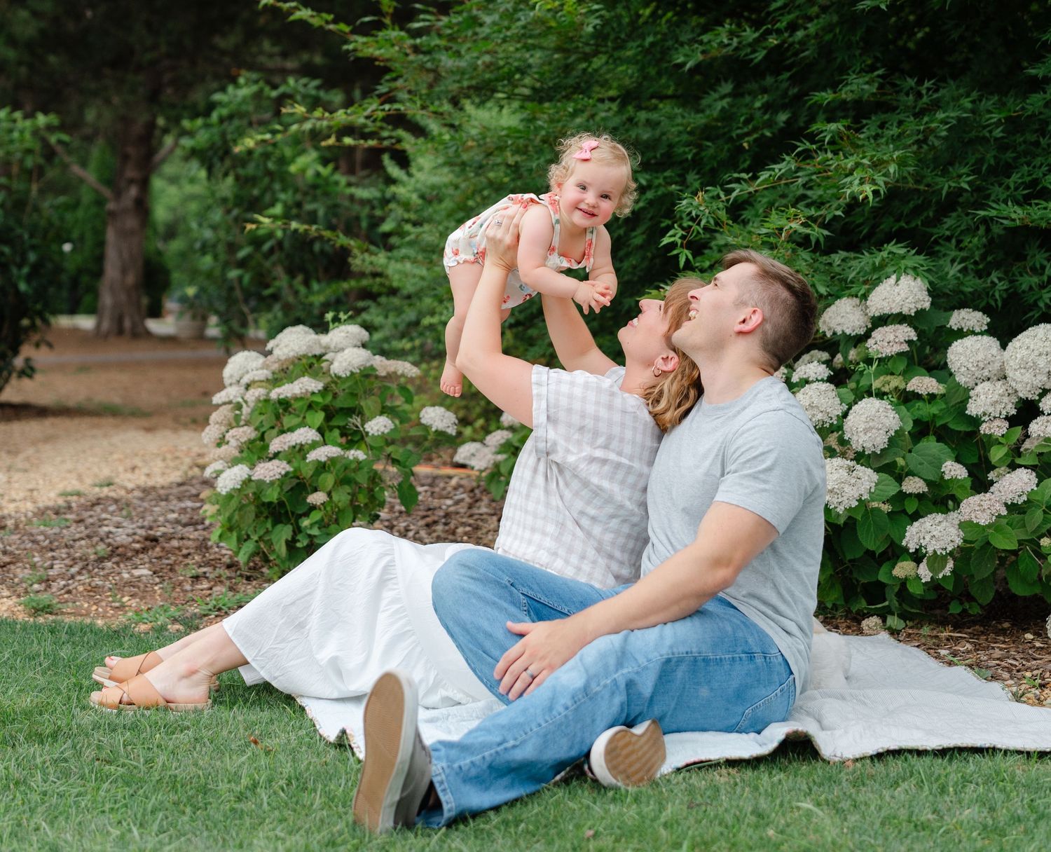 A happy family sits on a blanket in a lush garden, parents lifting their smiling baby in the air.