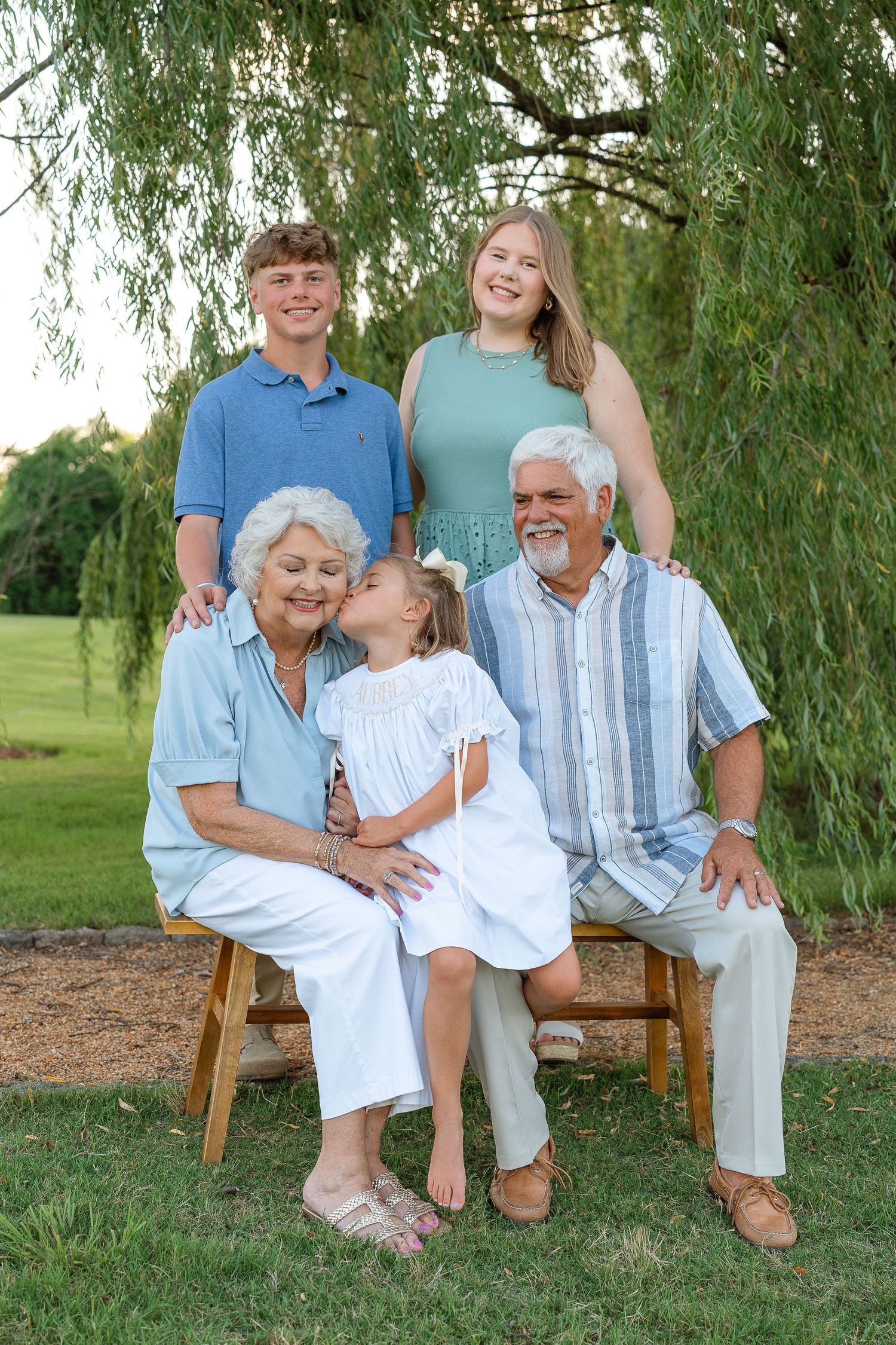 Grandparents seated on bench with three grandchildren under willow tree in green park.