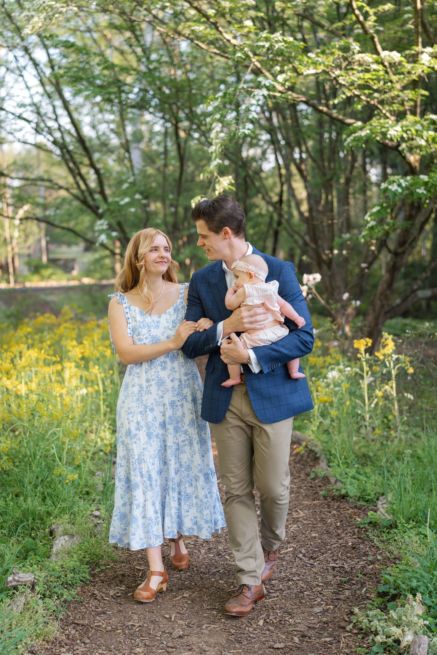 Couple in blue floral dress and navy blazer walks along a garden path holding their baby.