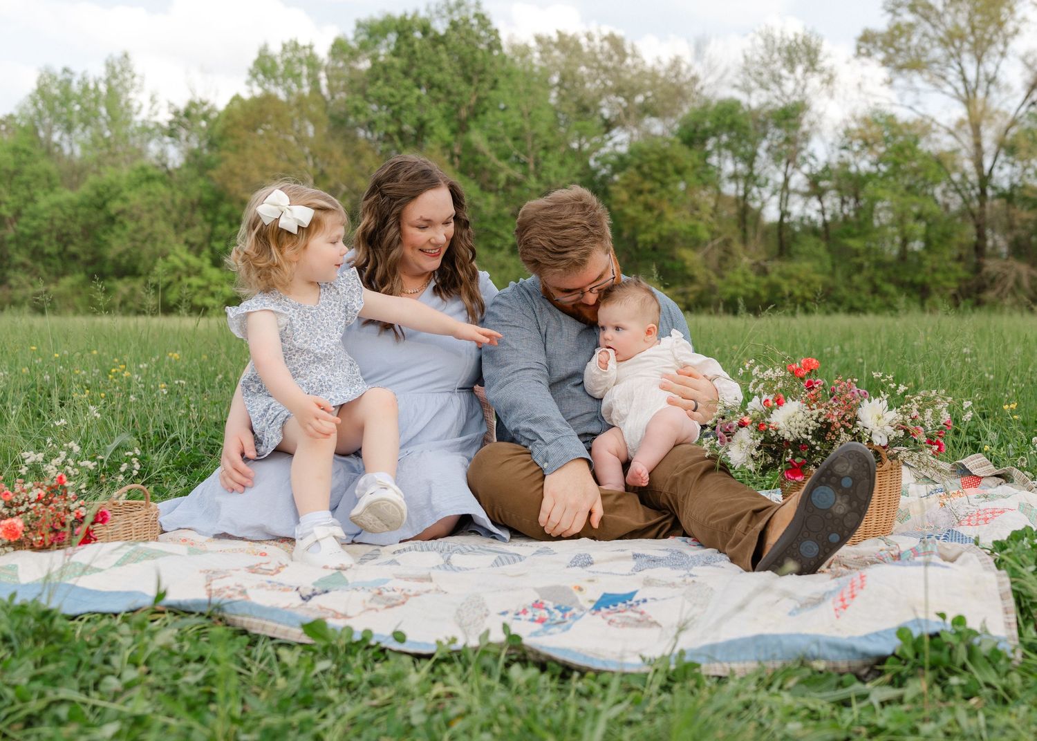A family of four relaxes on a floral quilt in a meadow, with parents lovingly attending to their children.