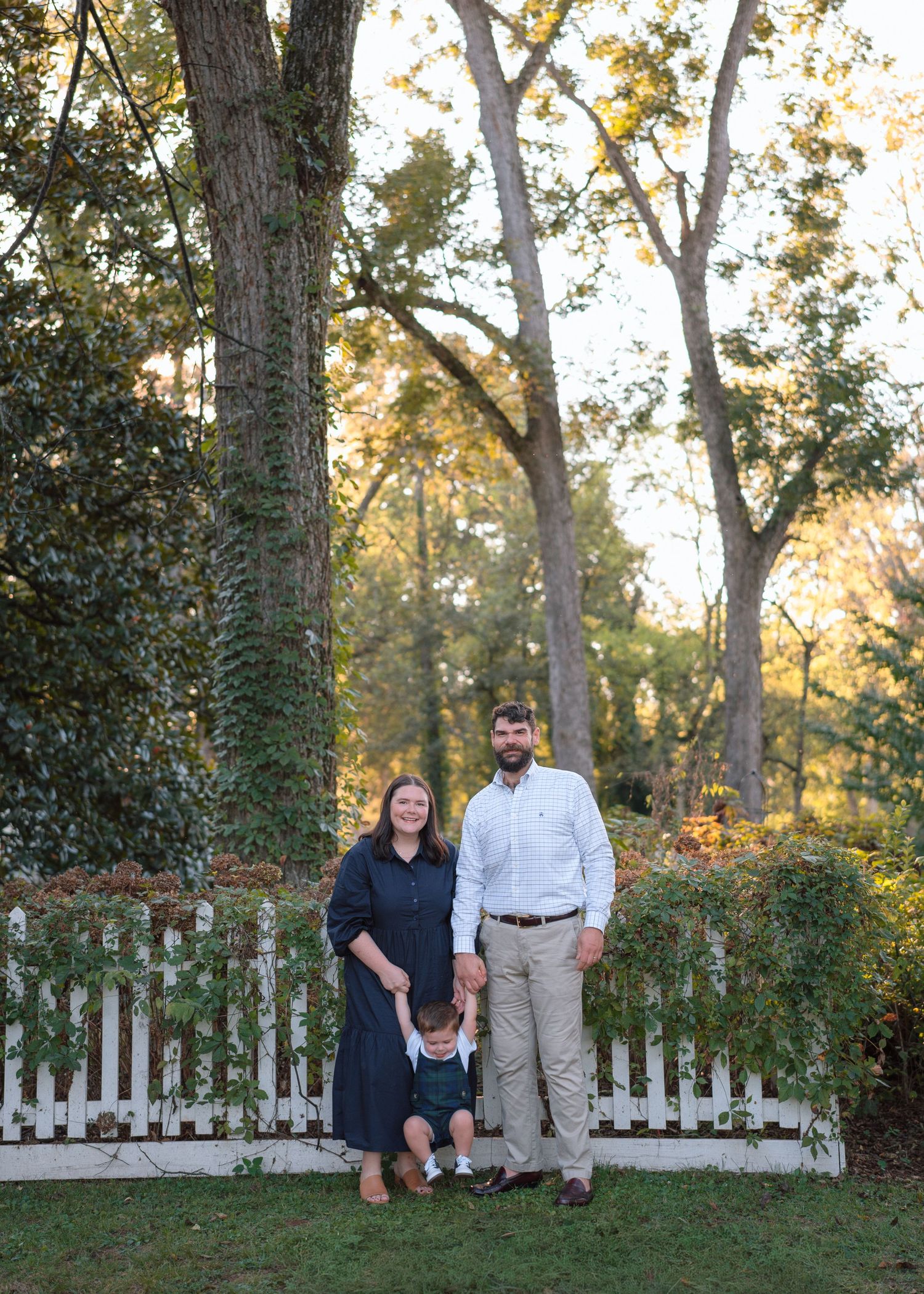 A family of three poses outdoors by a white picket fence surrounded by tall trees during golden hour.