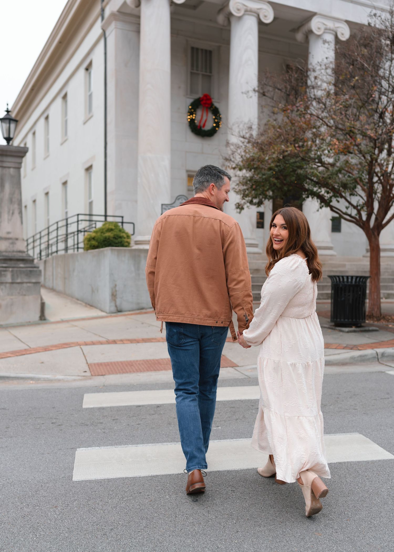 Happy couple holds hands crossing a street, woman in white dress looks back smiling, Christmas wreath on building behind.