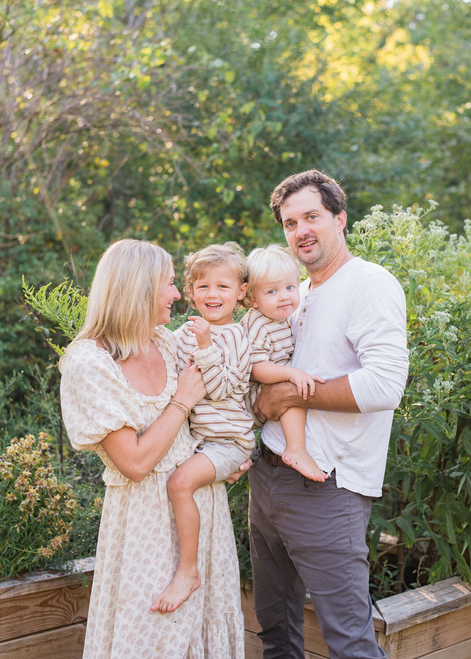 Joyful family of four with two young children smiling warmly together in a lush green outdoor setting.