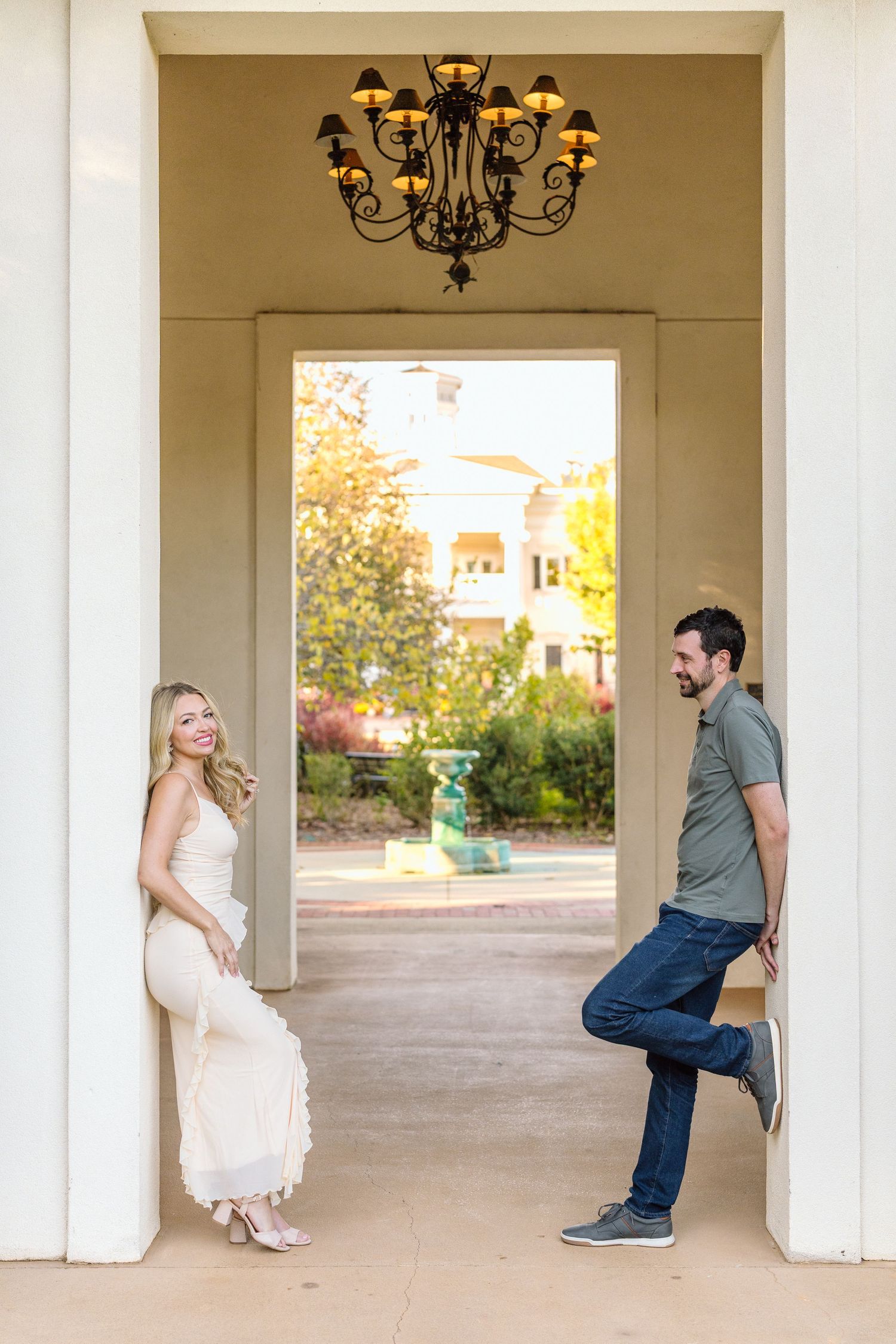 Engaged couple posing playfully on opposite columns of a grand white portico with chandelier above.