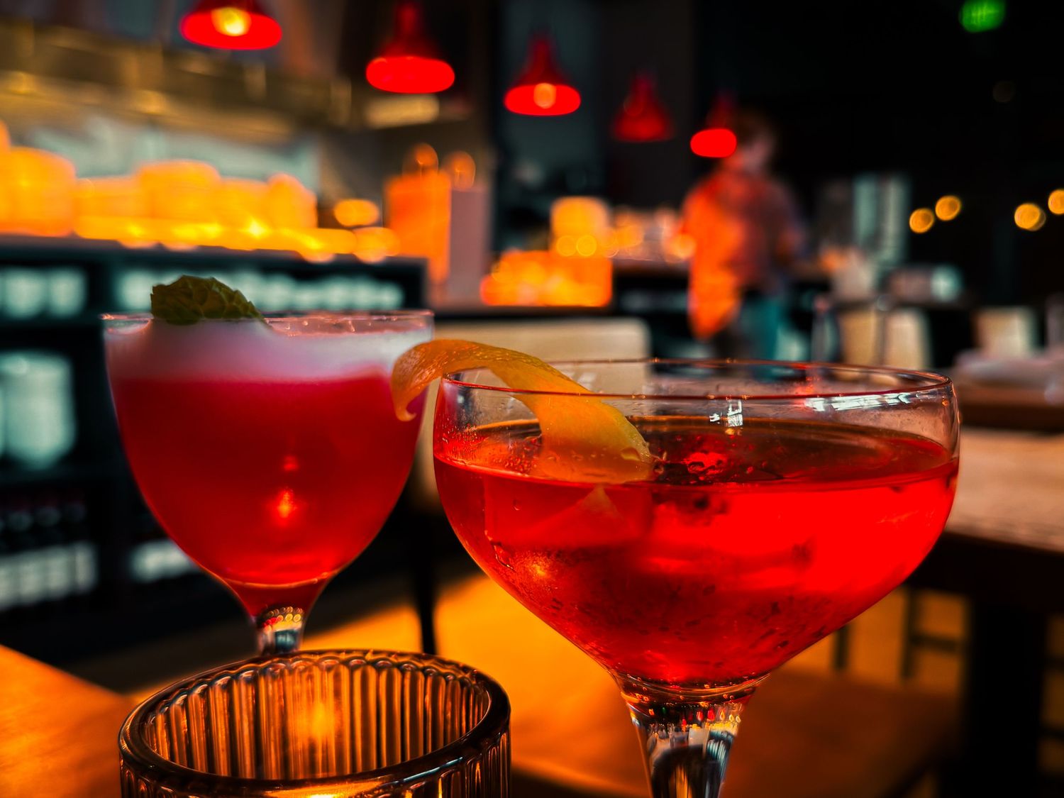 Two red cocktails garnished with orange slices in coupe glasses at a dimly lit bar.