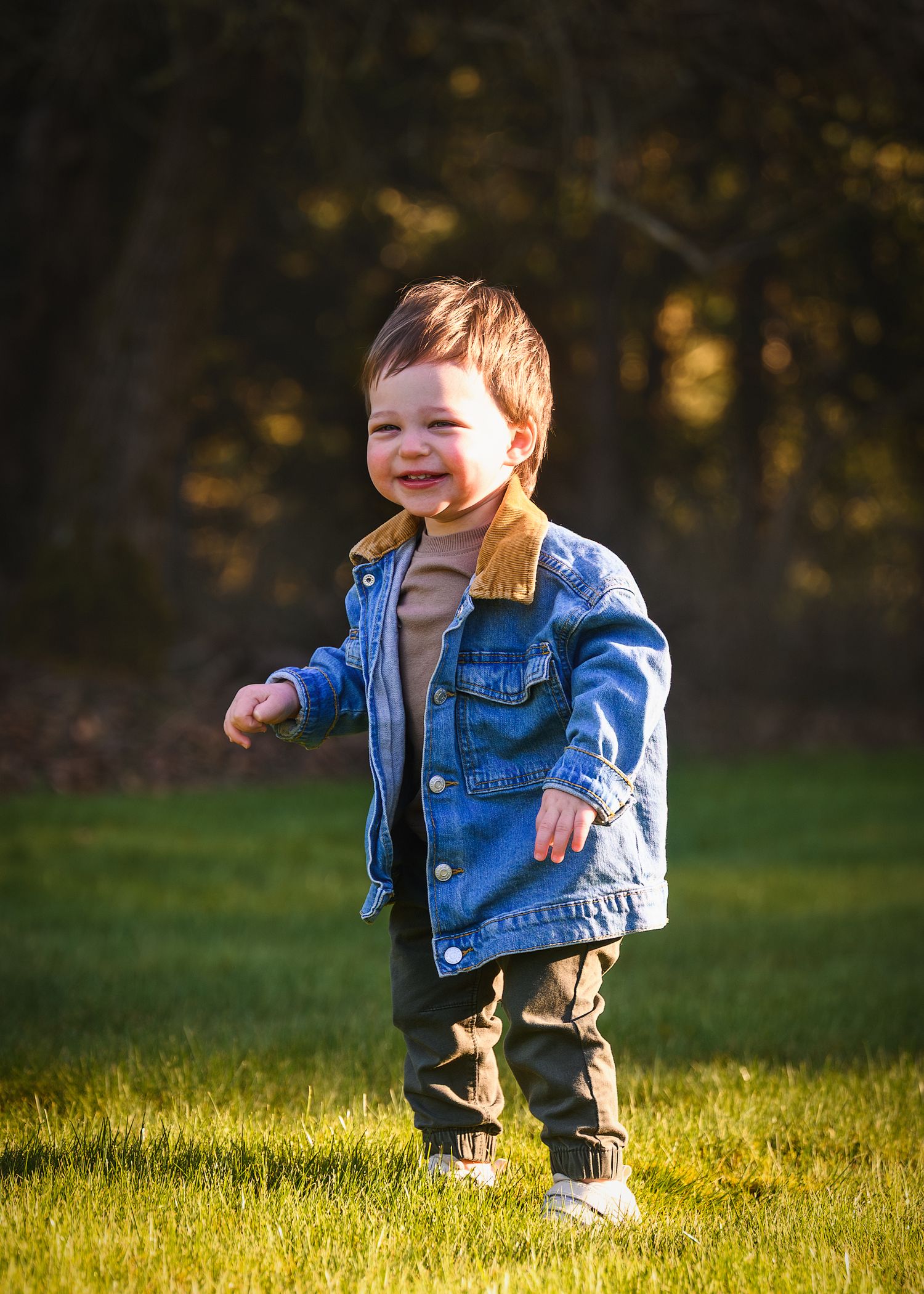 A young child in a denim jacket and khaki pants plays in a grassy field during golden hour sunlight.