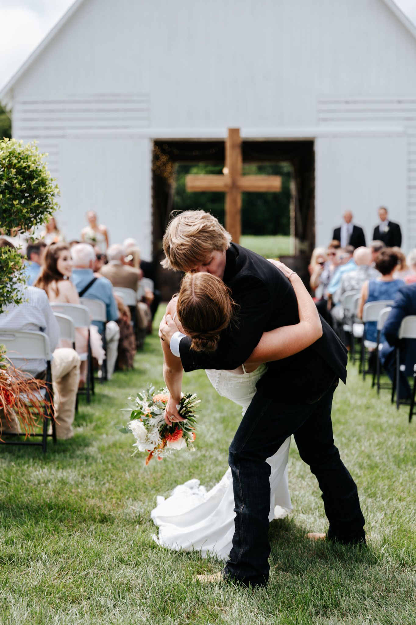 A romantic wedding kiss in front of a white church with guests seated on chairs during an outdoor ceremony.