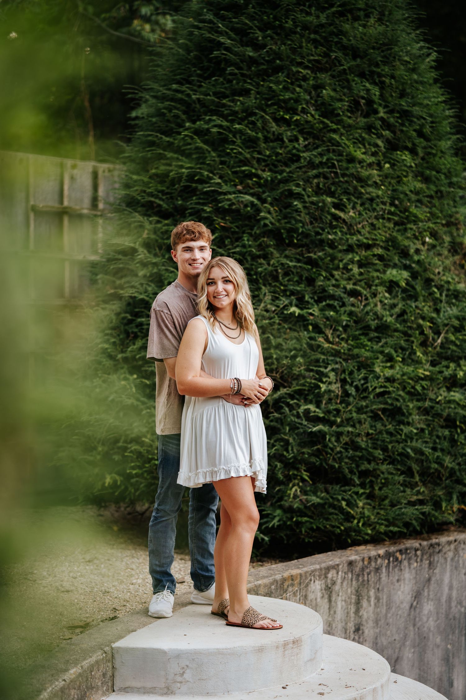 Romantic couple poses together on concrete ledge with green hedge background.