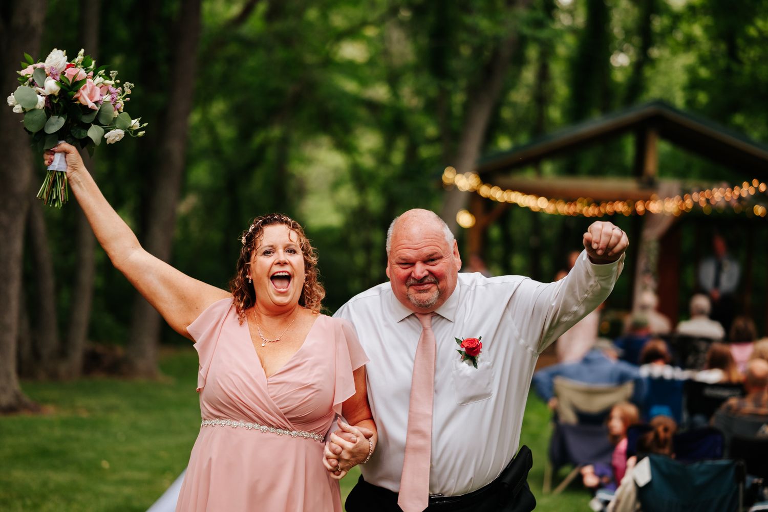 People celebrate joyfully at an outdoor wedding reception under trees.