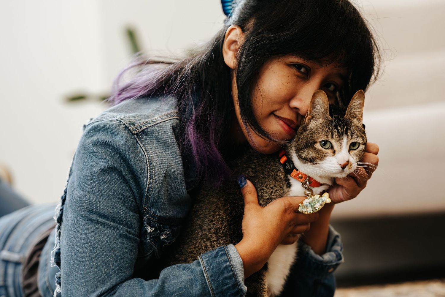 Person in denim jacket snuggling with a white and brown cat indoors.