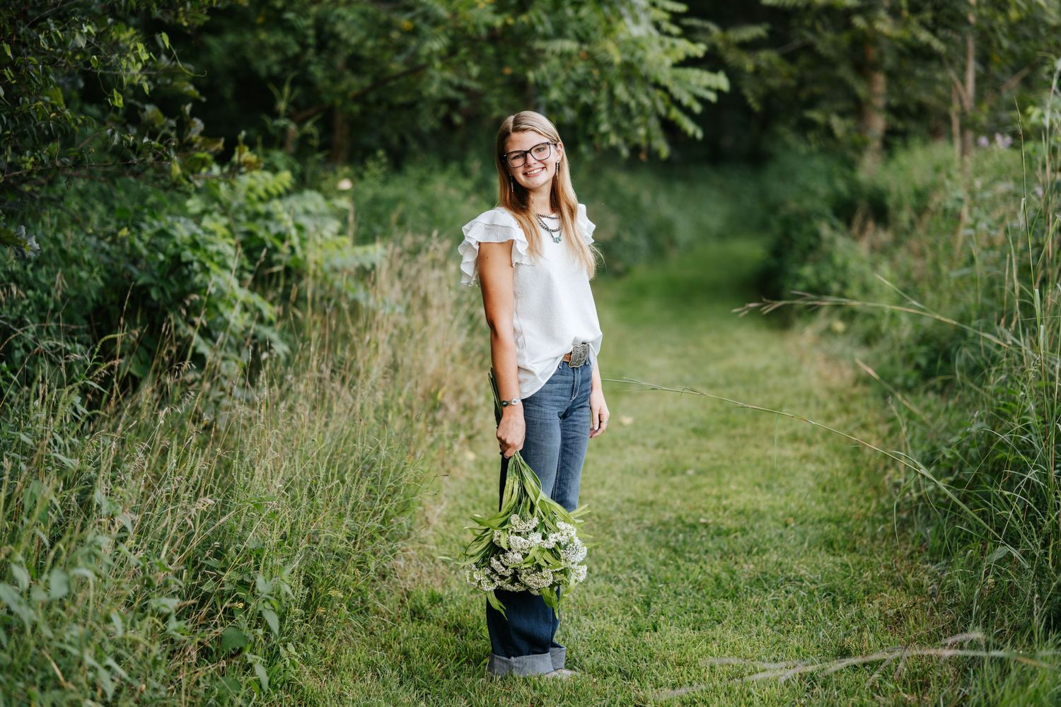 A person wearing jeans and a white t-shirt walks along a green nature path on a sunny summer day.