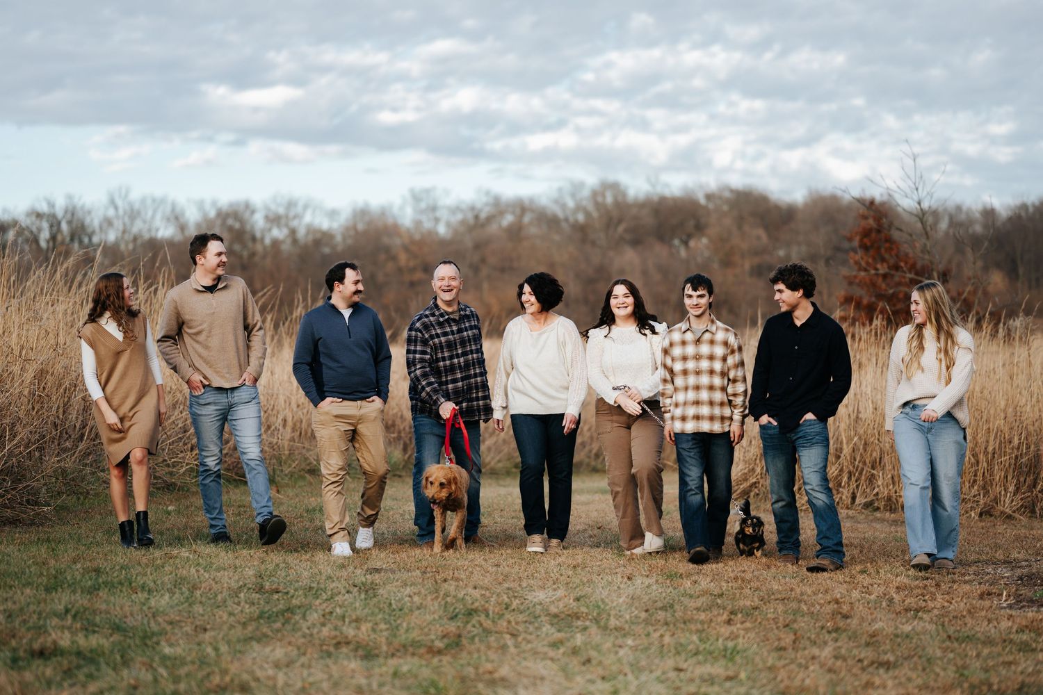 A large family group poses together in a field with tall golden grasses and cloudy skies in a casual outdoor portrait.