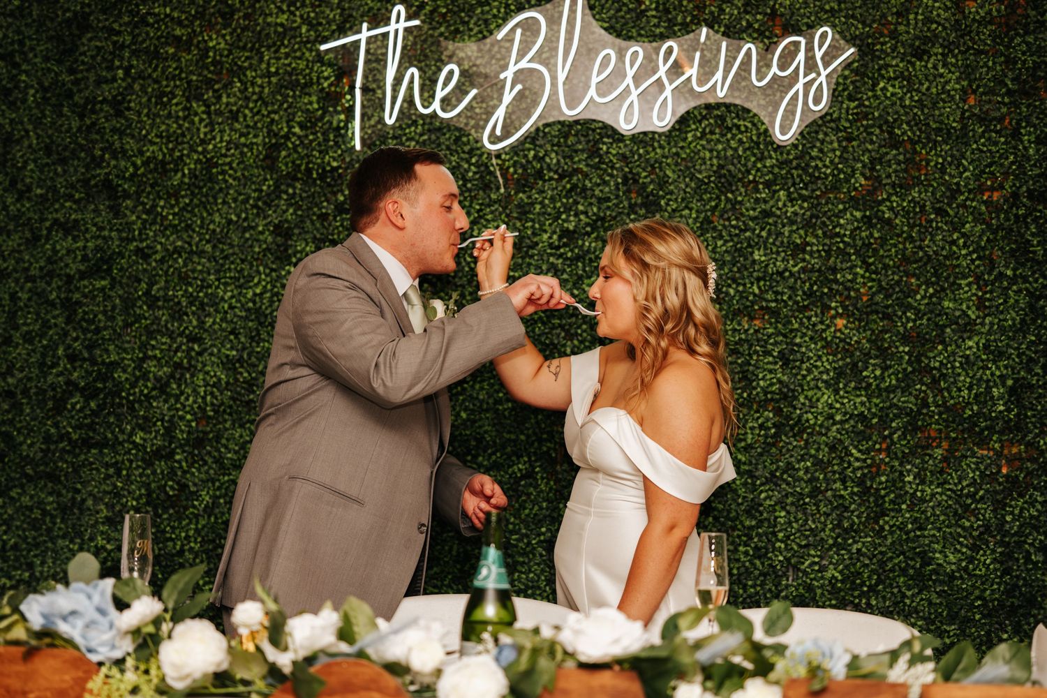 Wedding couple sharing a celebratory toast in front of a neon sign and greenery wall.