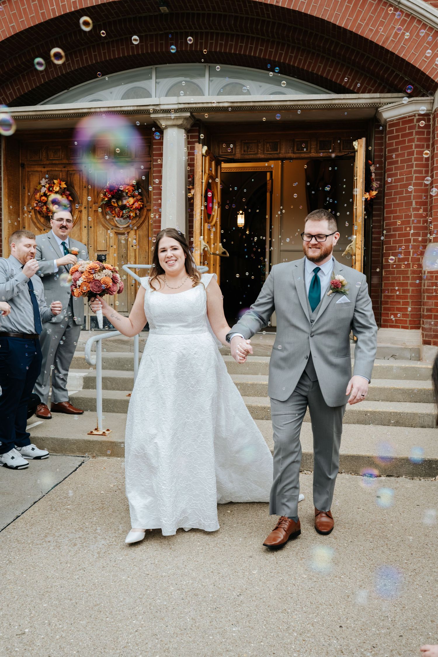 Newlyweds walking outside brick church entrance with bubbles floating around them.