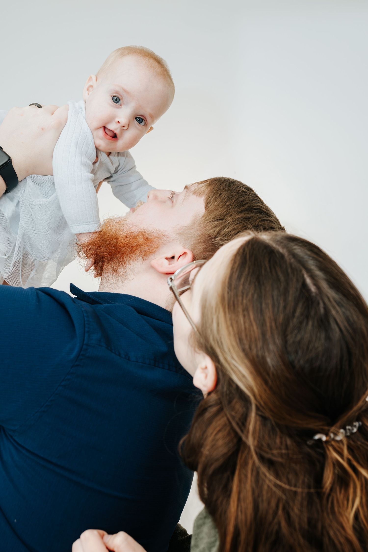 Parents lift and play with their baby wearing a light gray onesie against a white background.