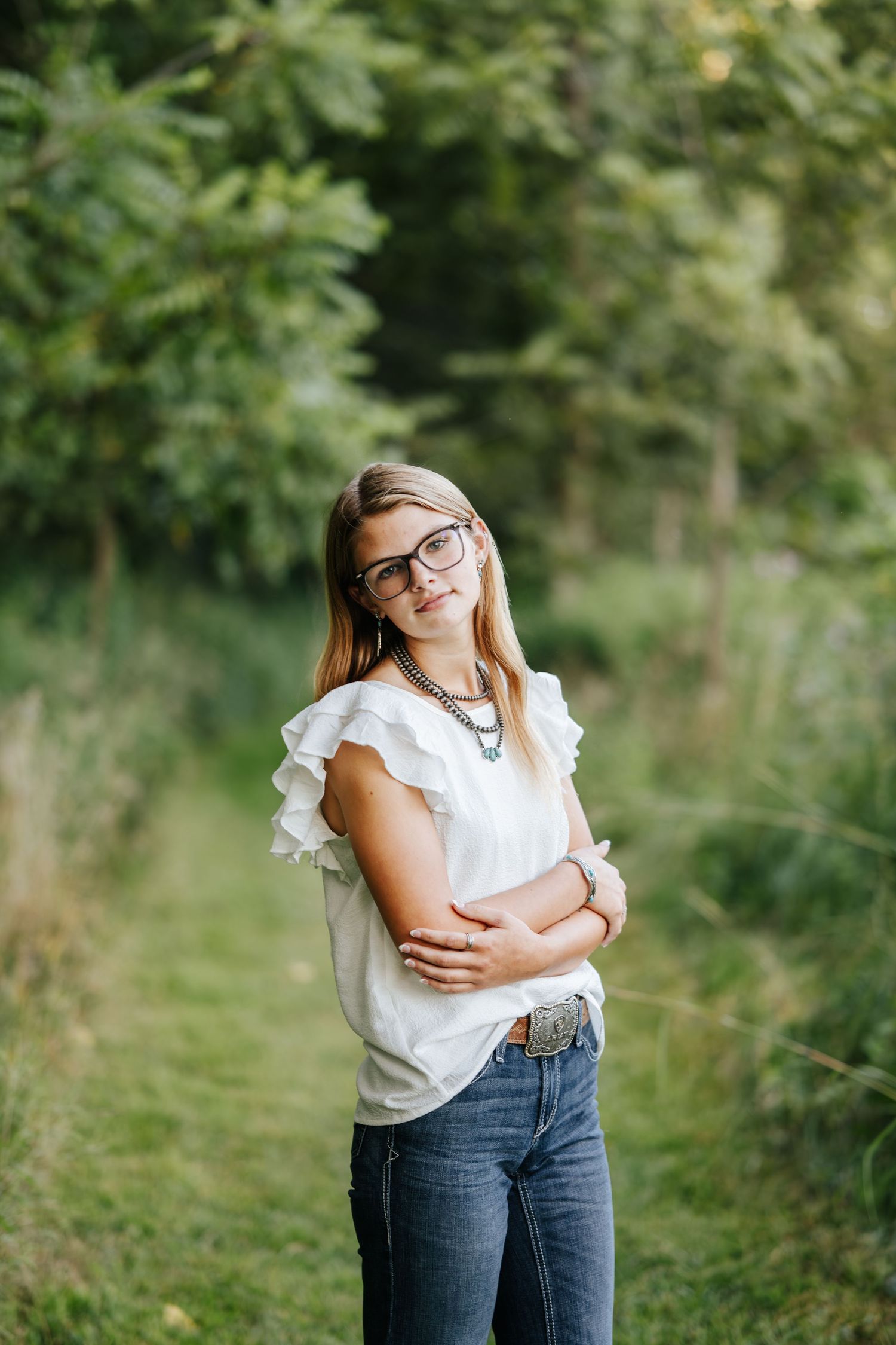 Person in white ruffled top and jeans poses playfully in a green outdoor setting.