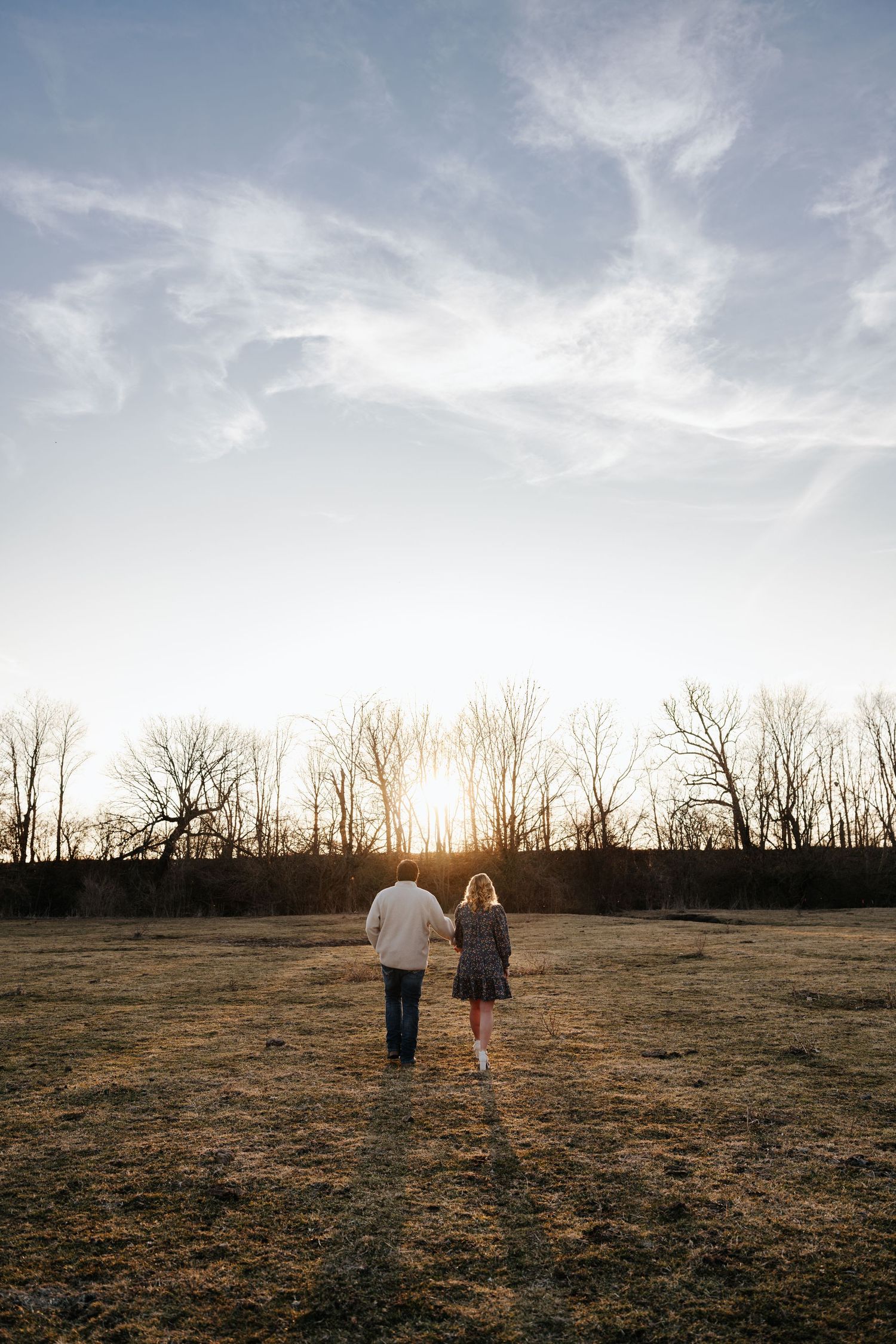 Two people walk together across a bare field at sunset with silhouetted trees in the background and cloudy skies above.