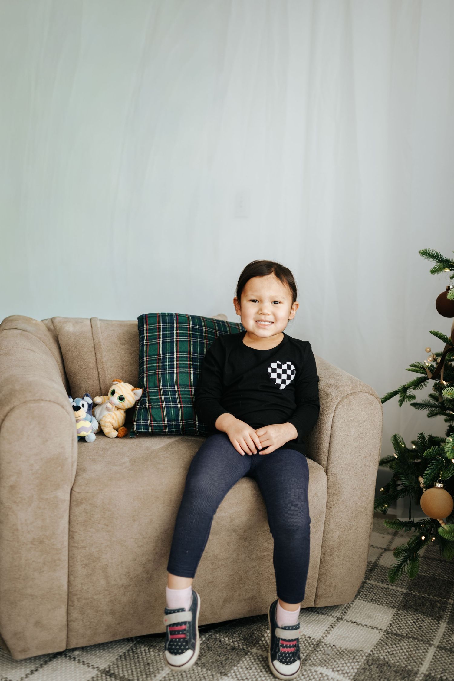 A small dog peeks out from behind a plaid pillow on a beige couch while someone sits nearby.