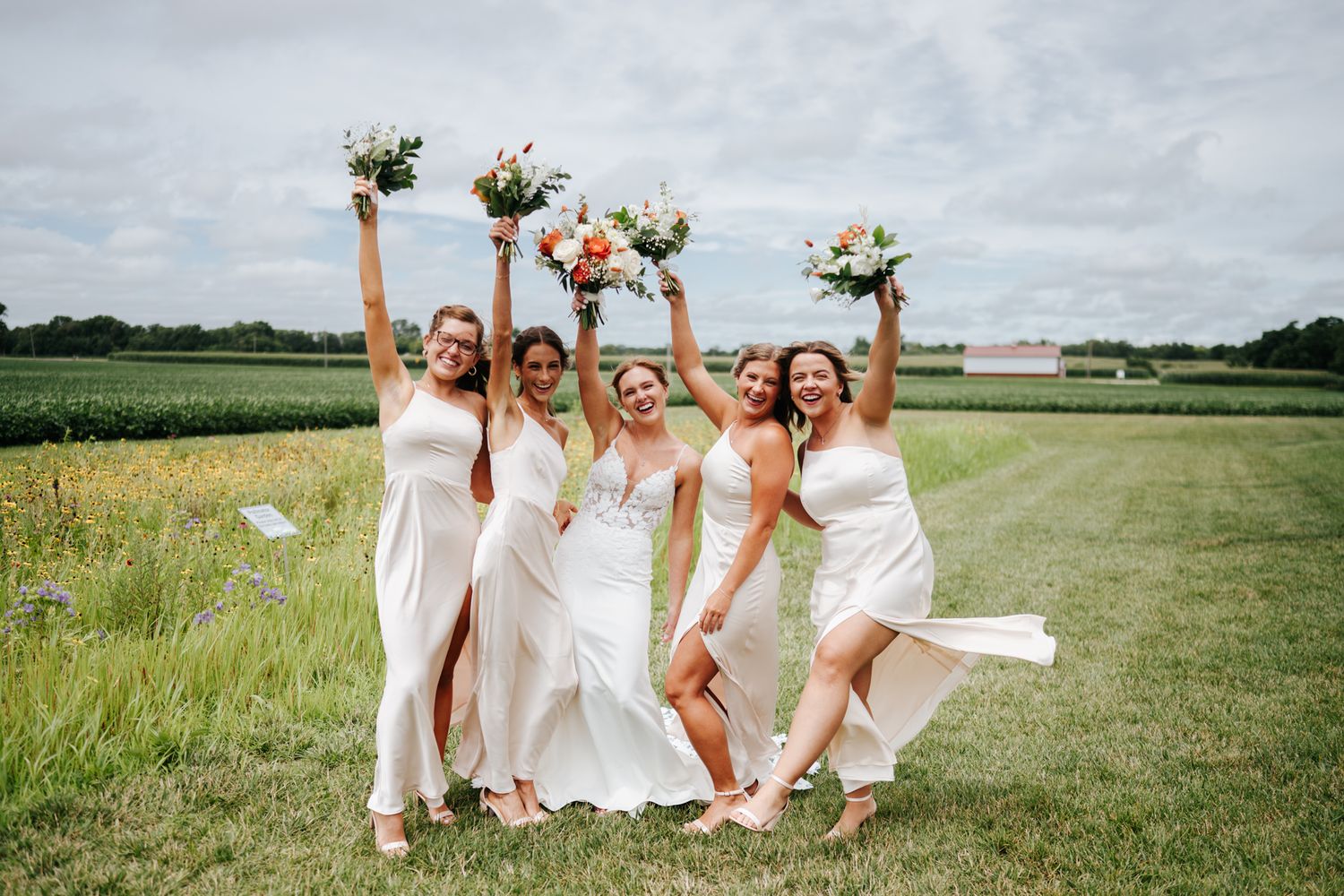 A bridal party celebrates in white dresses on a sunny field.