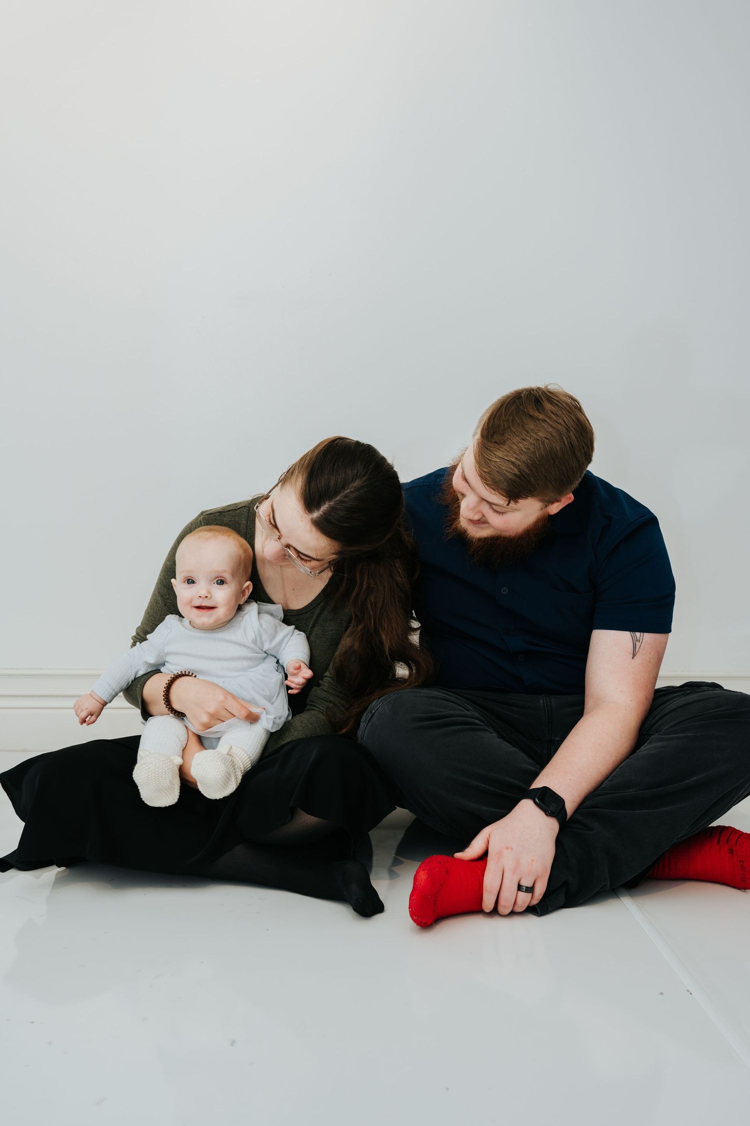 A family sits together on a white floor, sharing an intimate moment with their baby in a minimalist setting.