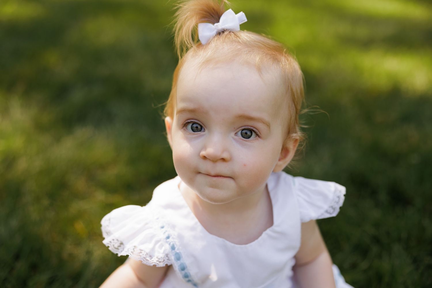 Adorable red-haired baby girl in white dress sitting on green grass, looking curiously at camera.
