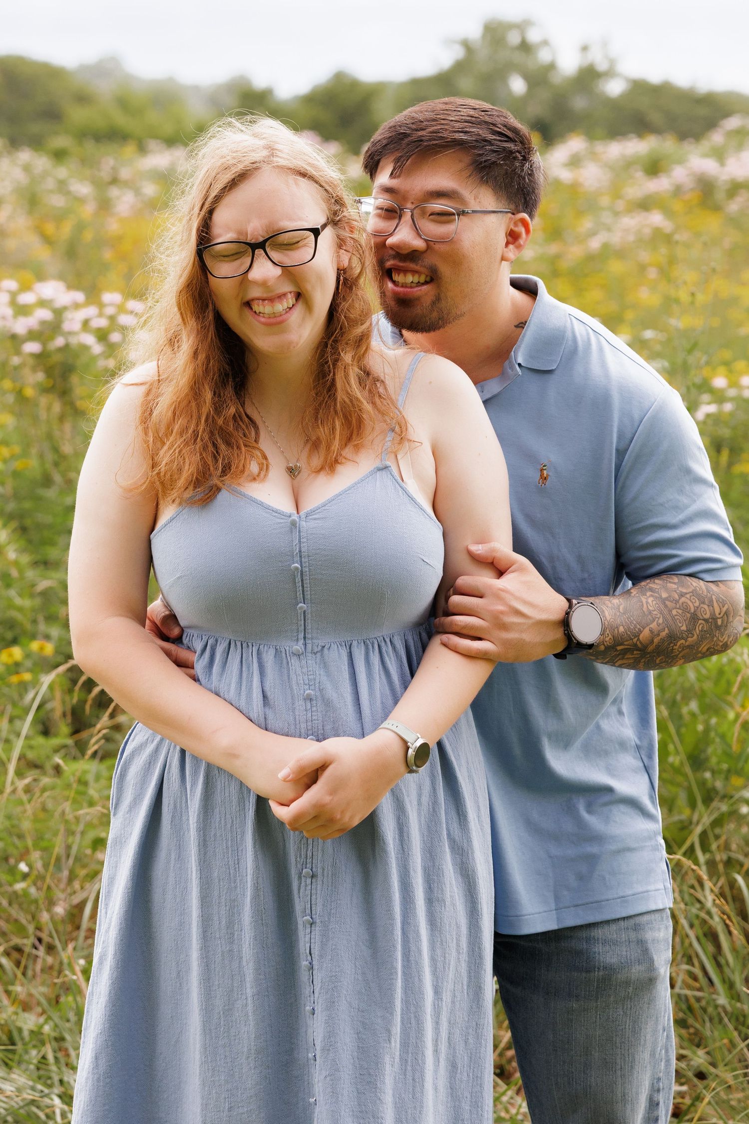 A smiling couple in blue outfits poses together in a wildflower field, the man embracing the woman from behind.