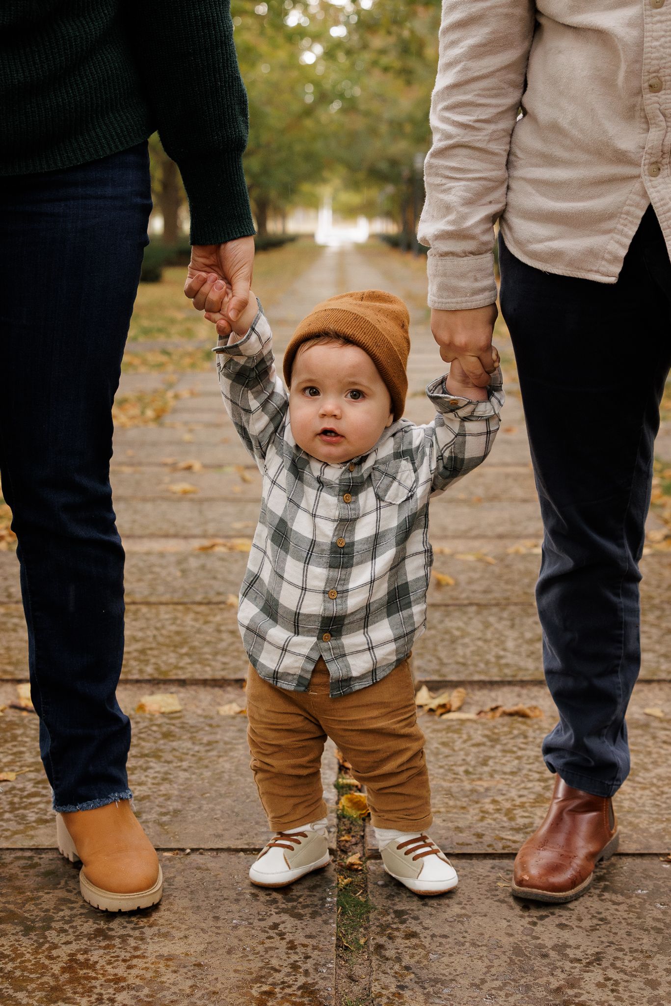 Toddler in plaid shirt and brown beanie holds parents hands on a tree-lined path in autumn.