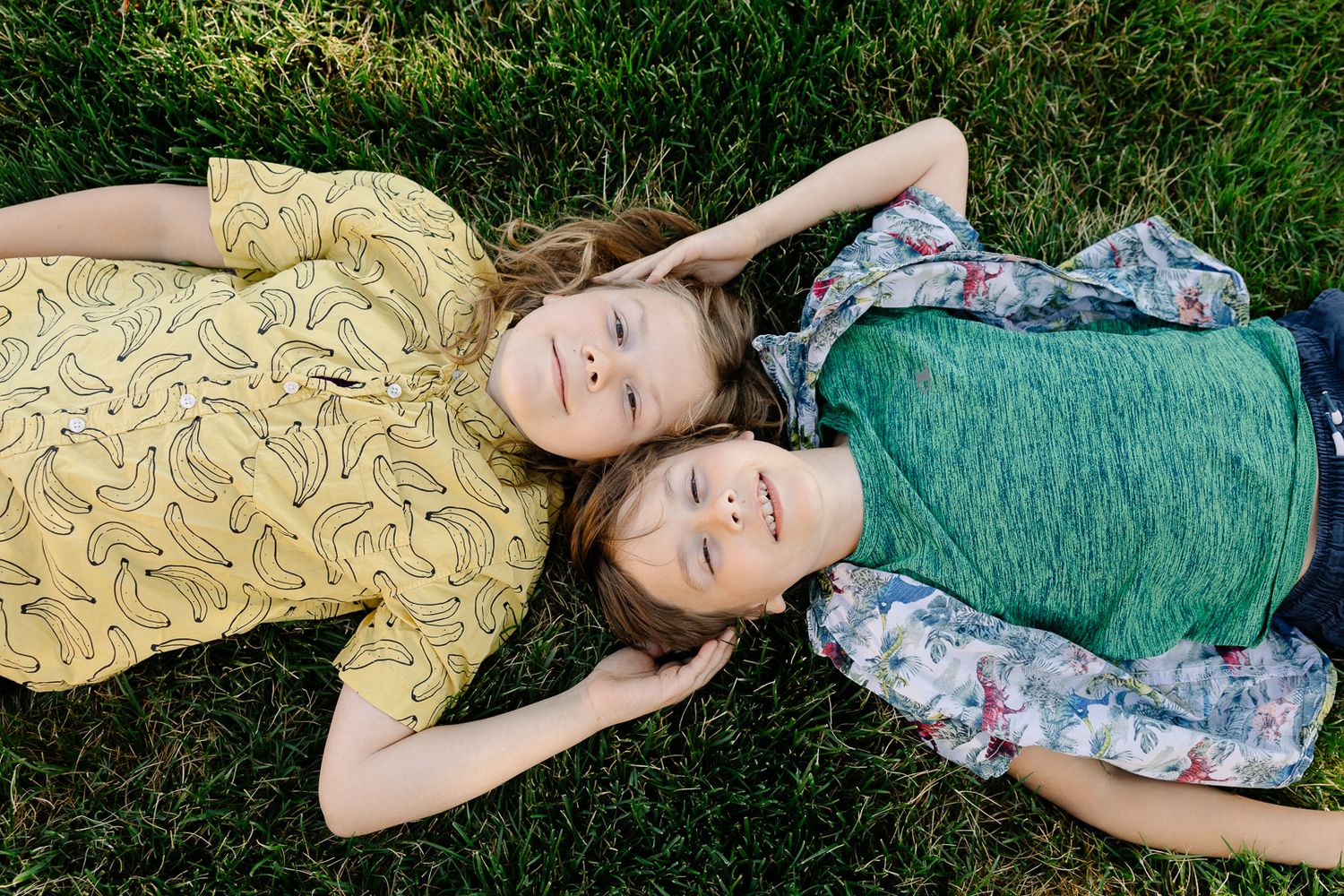 Two children lying on green grass, smiling up at camera, wearing colorful summer clothes.