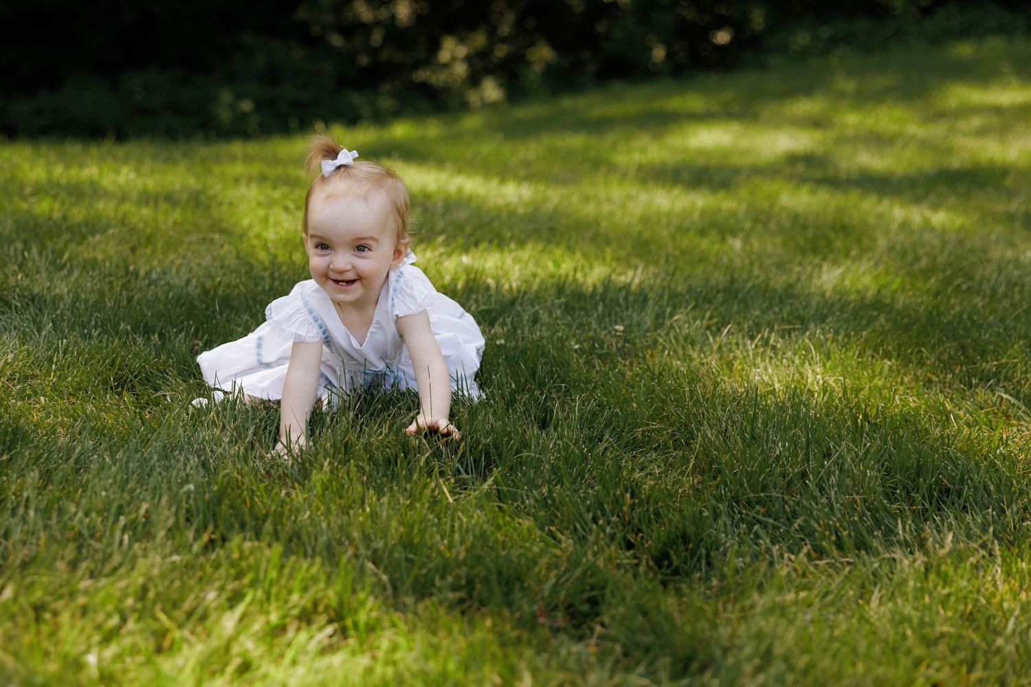 A happy baby in white outfit crawls on lush green grass outdoors in sunlight.