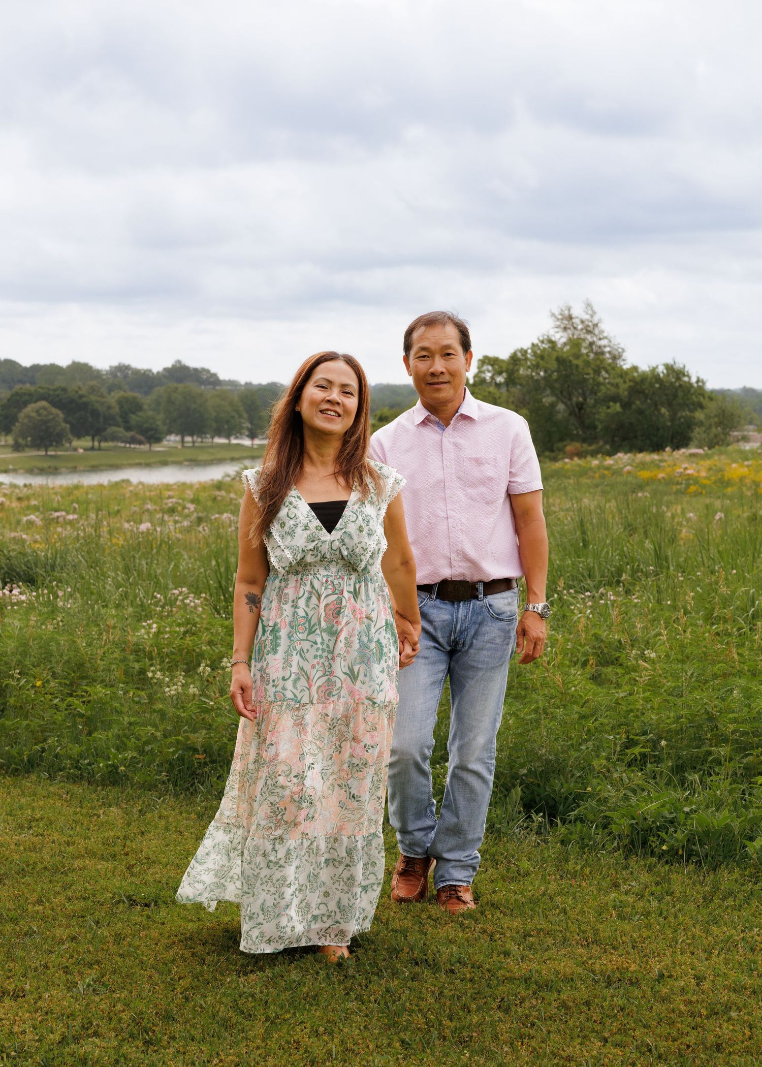 A couple walks hand-in-hand through a lush green meadow near a lake, smiling on a cloudy day.