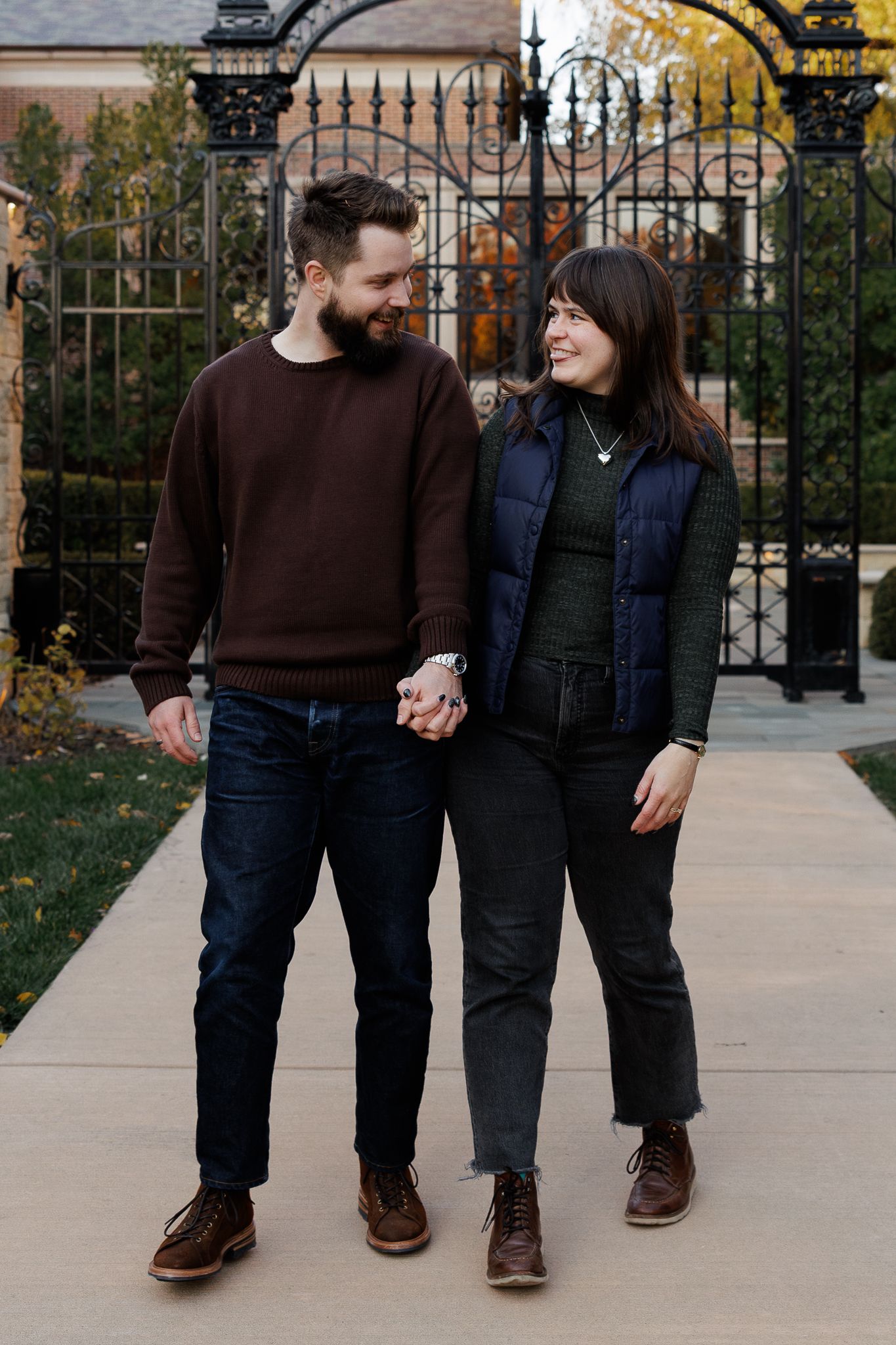 A smiling couple holds hands while walking on a pathway in front of decorative iron gates during autumn.