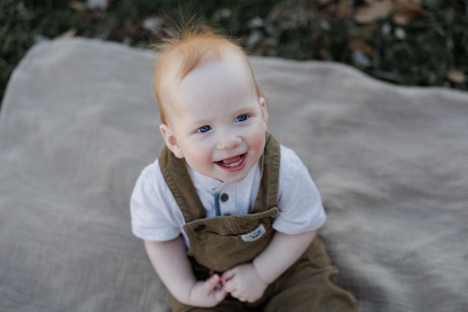 Smiling red-haired baby boy in white shirt and olive overalls sitting on a tan blanket outdoors.