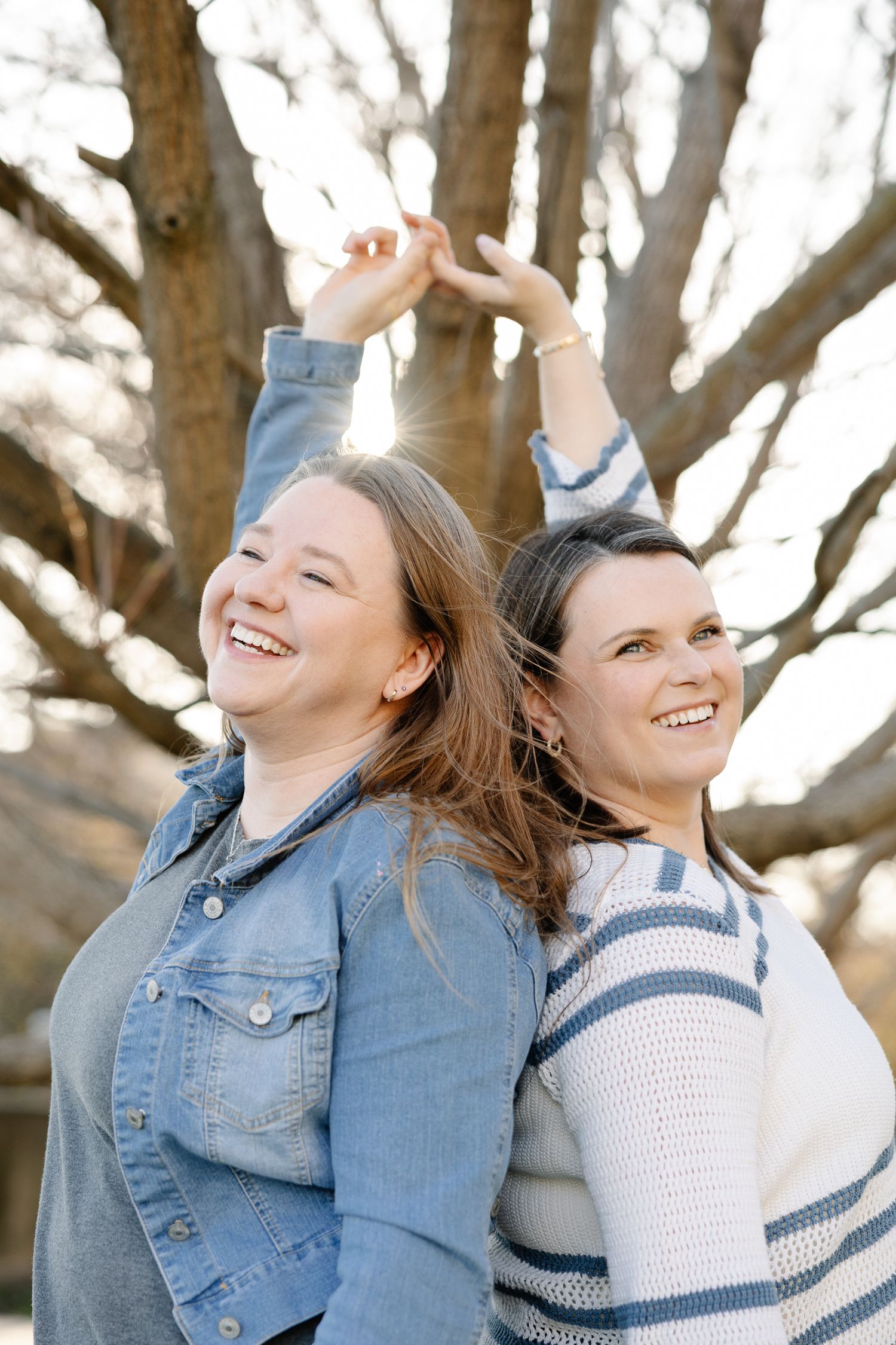 Two laughing women stand back-to-back outdoors, holding hands raised above, with a large tree in the background.