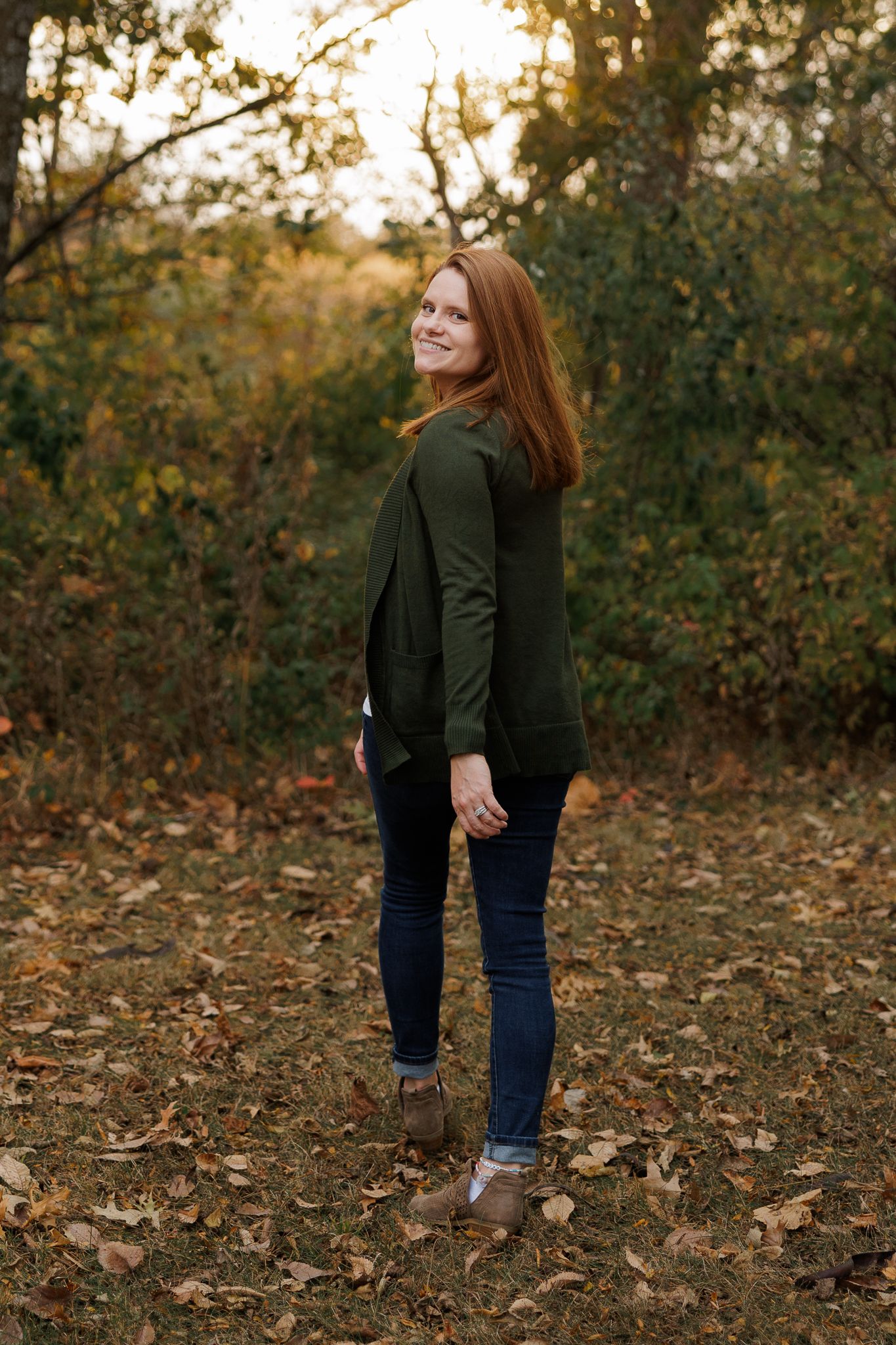 A smiling redhead woman in green top and jeans stands among autumn leaves in a wooded outdoor setting.