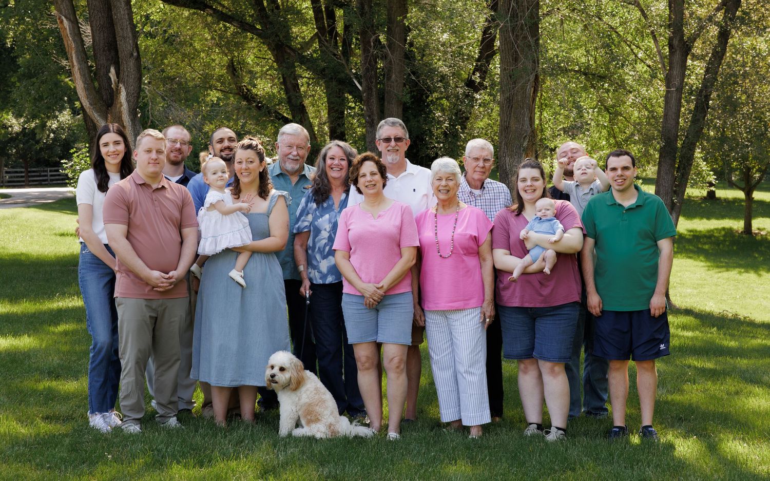 Large family group portrait outdoors on green grass with trees, including adults, children, and a fluffy white dog.