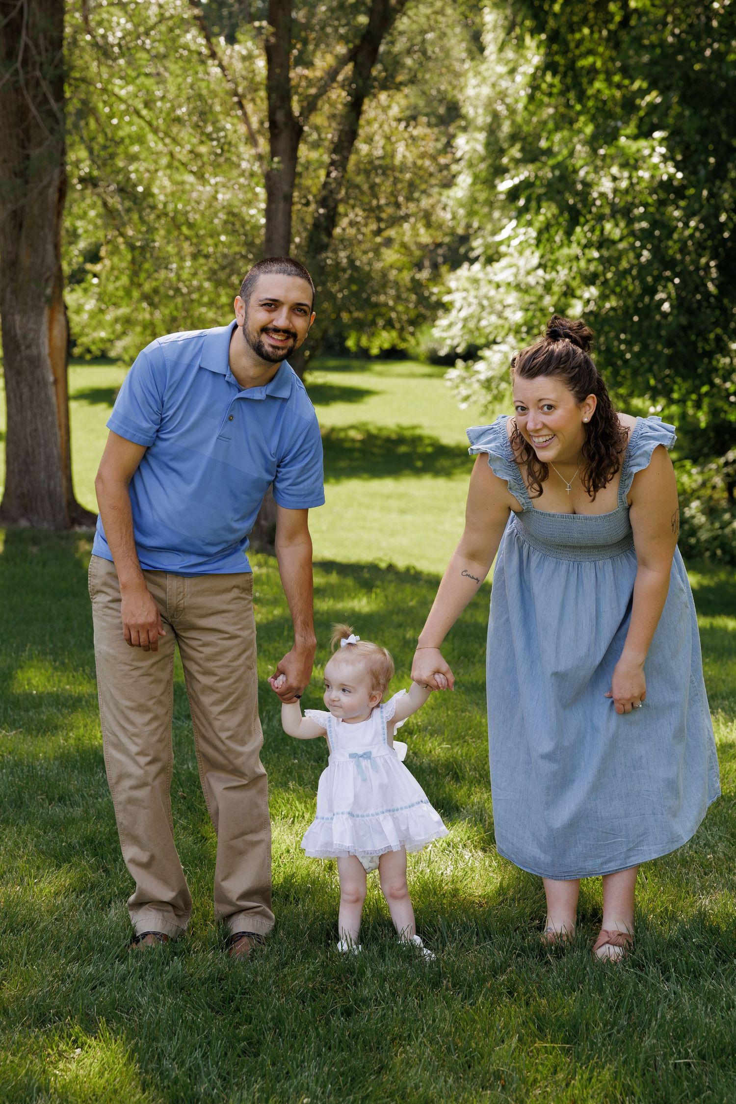 A happy family with a man in blue polo, woman in blue dress, and toddler girl in white dress holding hands in a green park.