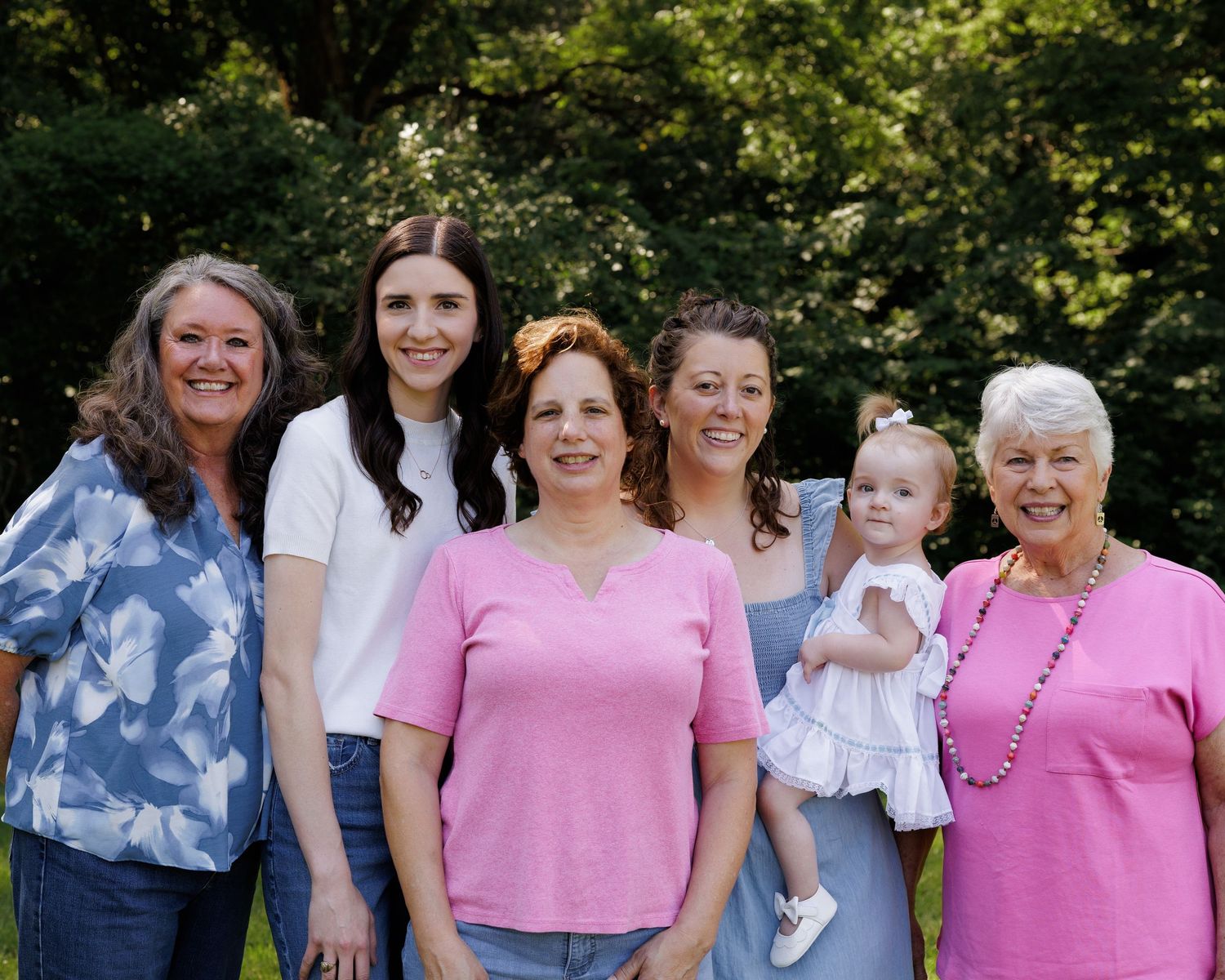 Six women of different generations smile together outdoors, including a grandmother, mothers, and a baby girl in a white dress.