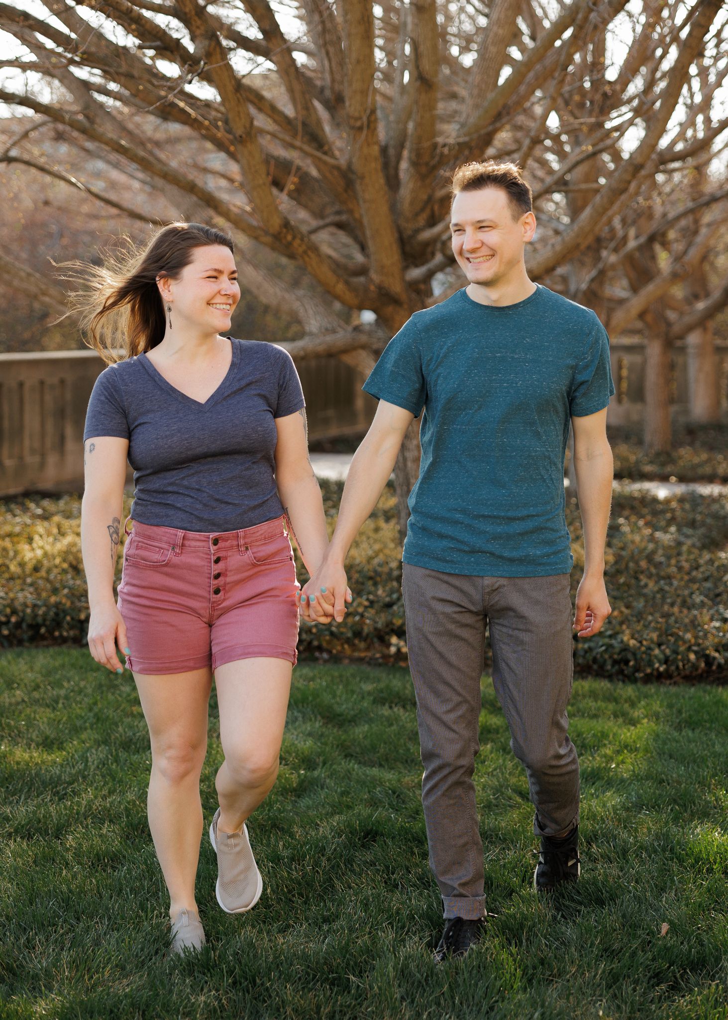 A smiling couple holds hands while walking on green grass outdoors, with bare trees in the background.